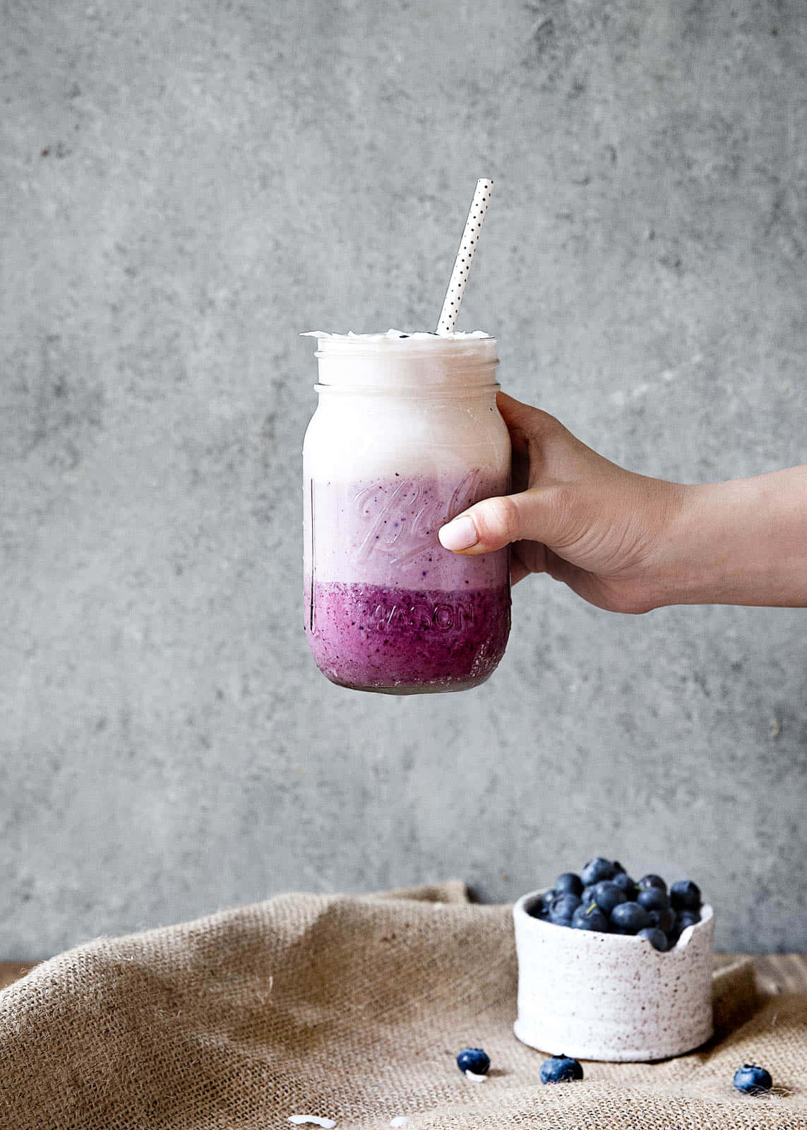 Blueberry Smoothie 桌布 壁紙 — 一隻手拿著一個裝在玻璃瓶中的分層藍莓奶昔，背景是帶有紋理的灰色牆壁，旁邊有新鮮藍莓。