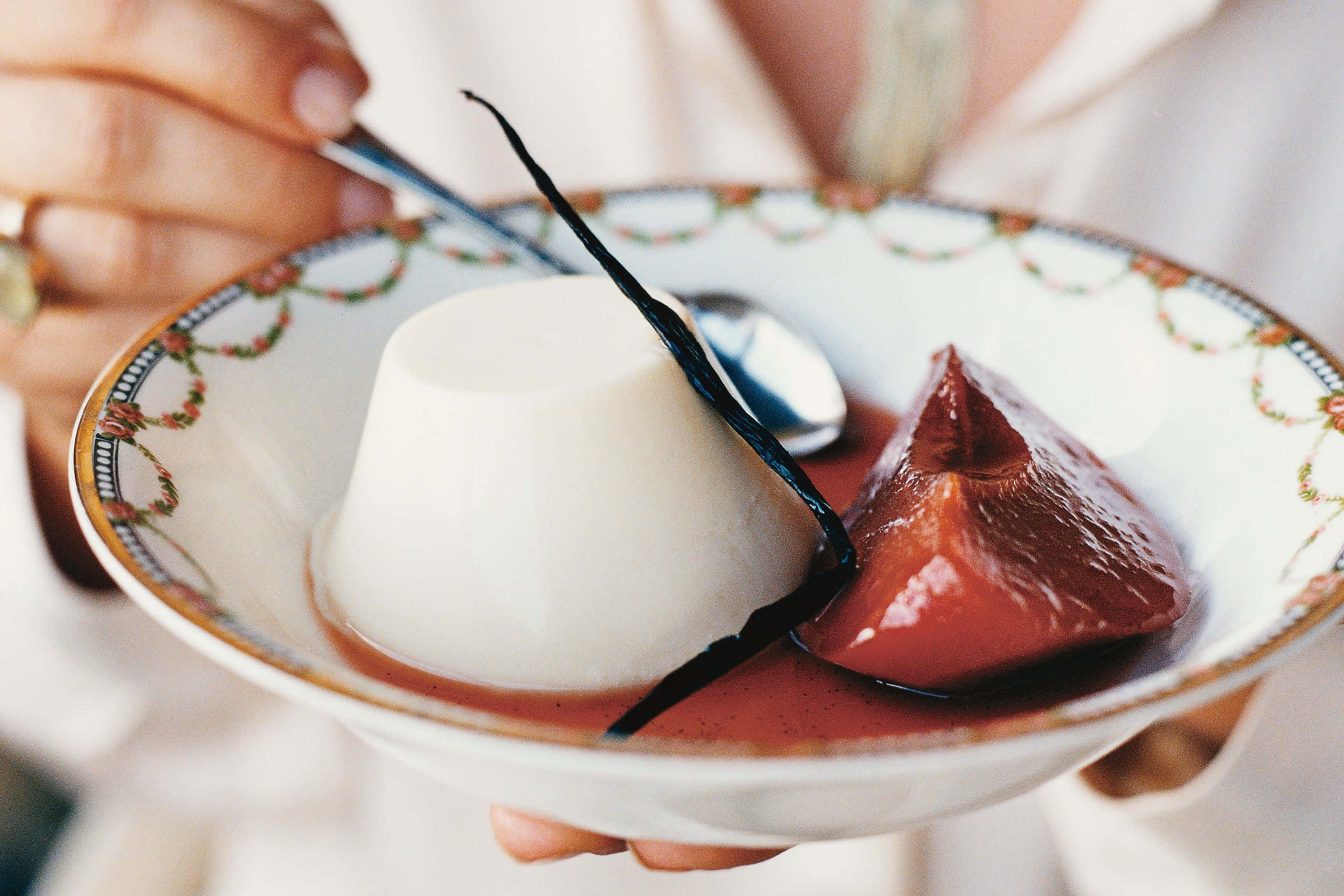 Blancmange 桌布 壁紙 — 一隻手拿著一個裝飾碗，裡面有奶油白色的意式奶凍和光澤的深紅色水果蜜餞，並用香草豆裝飾。