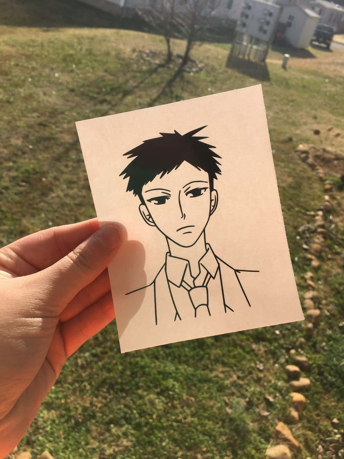 Takashi Morinozuka 桌布 壁紙 — 一隻手拿著一幅黑色墨水線條畫，畫中是一個動漫風格的男性角色，有尖刺頭髮和帶領的襯衫，背景是陽光普照的草地戶外環境。