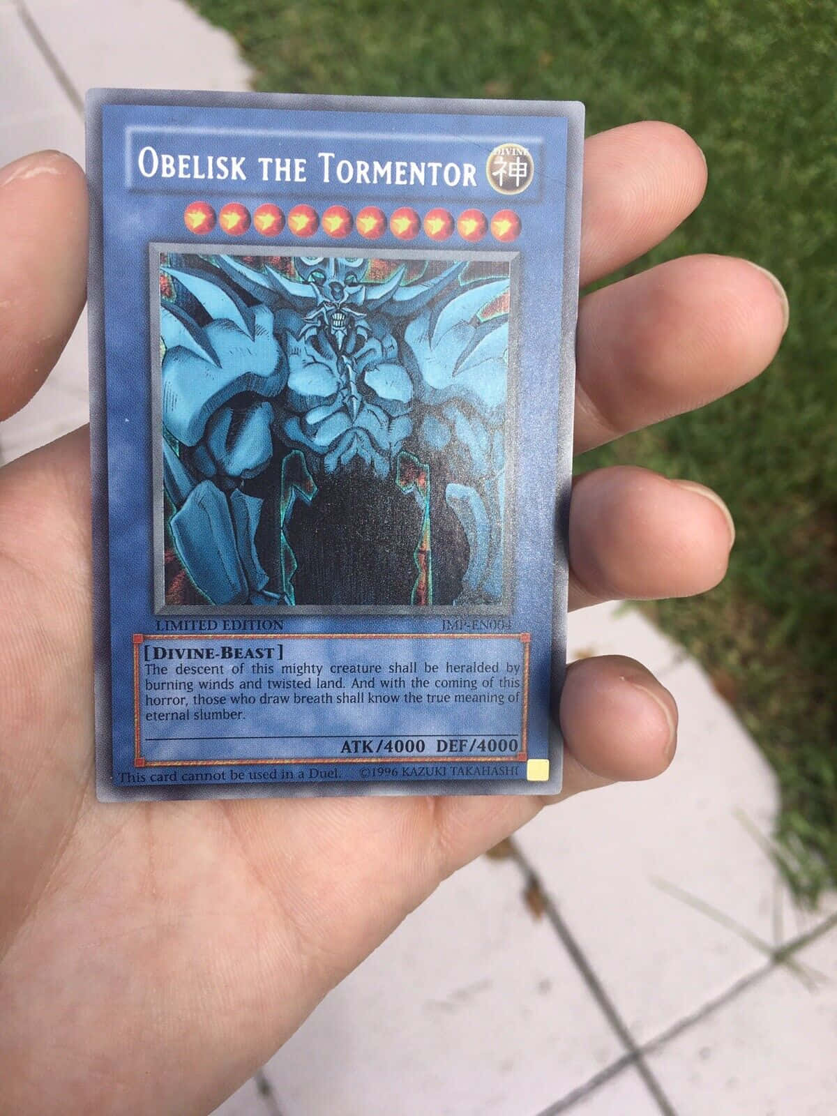 Obelisk The Tormentor 桌布 壁紙 — 一隻手拿著一張藍色限量版遊戲王歐貝利斯克的巨神兵卡，背景是草地戶外。