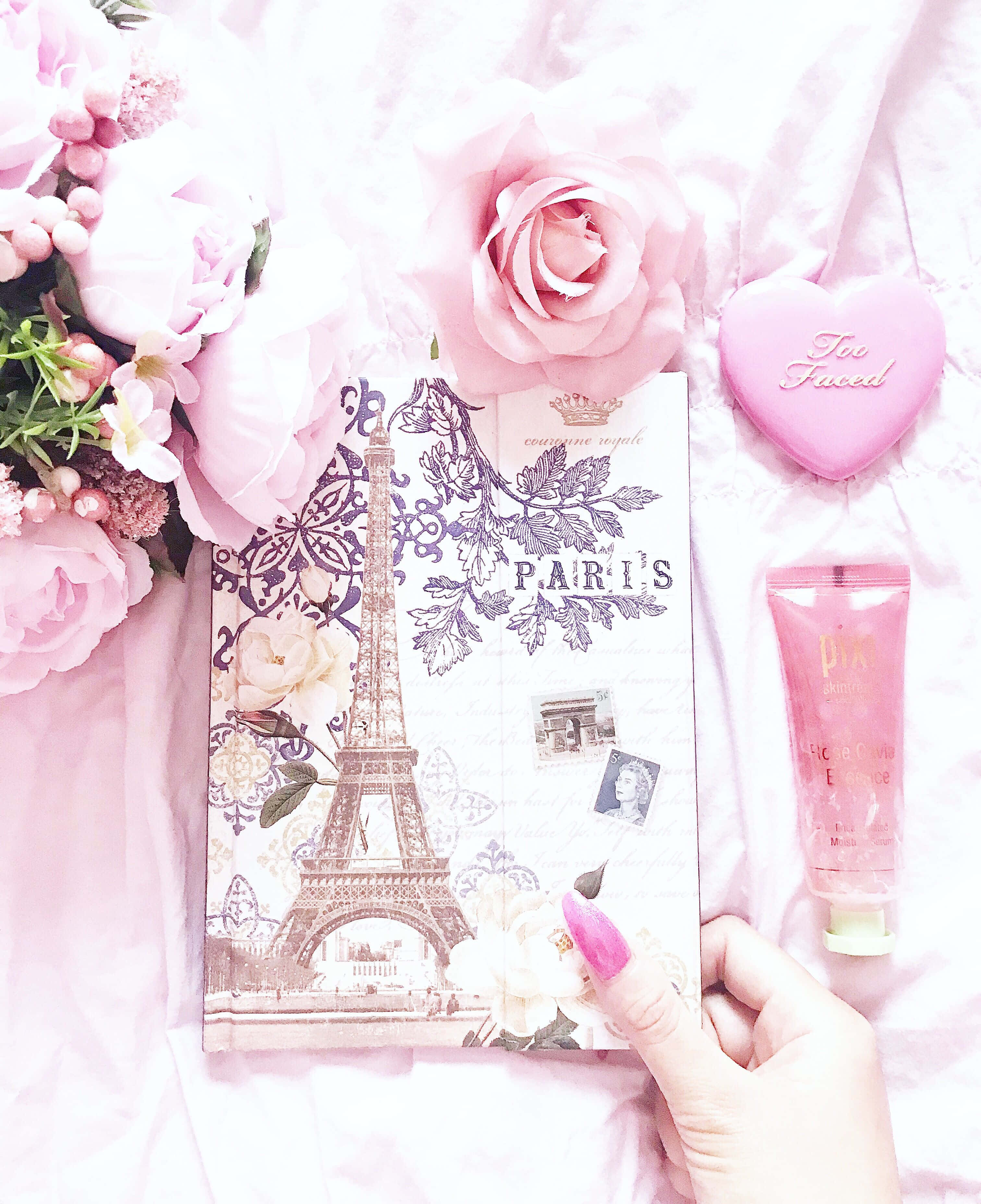 Eiffel Tower 桌布 壁紙 — 一隻手拿著一本巴黎主題的筆記本，周圍環繞著粉紅玫瑰和美容產品，放在柔軟的粉紅布料上。