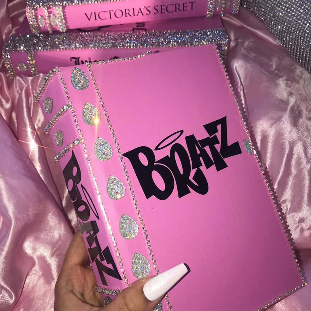 Baddie Vintage 90's Aesthetic 桌布 壁紙 — 一隻手拿著一本閃粉覆蓋的粉紅色 Bratz 書，帶有 Victoria's Secret 品牌標誌，鑲嵌在緞布中。