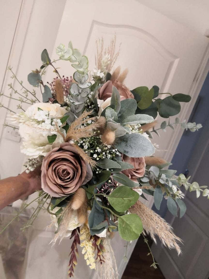 Roses Wedding 桌布 壁紙 — 一隻手拿著一束由塵粉玫瑰、桉樹和蒲葦組成的茂盛花束，背景是柔和的白色門。
