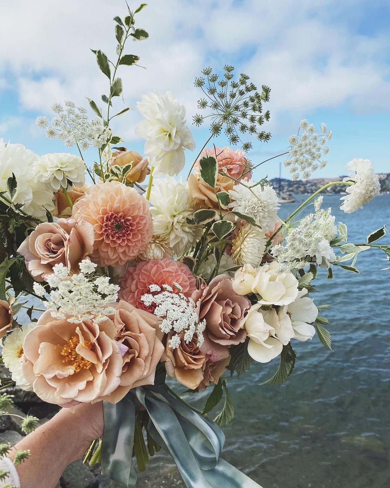 Roses Wedding 桌布 壁紙 — 一隻手拿著一束由蜜桃白和奶油色花朵組成的豐盛花束，背景是藍色的水和天空。