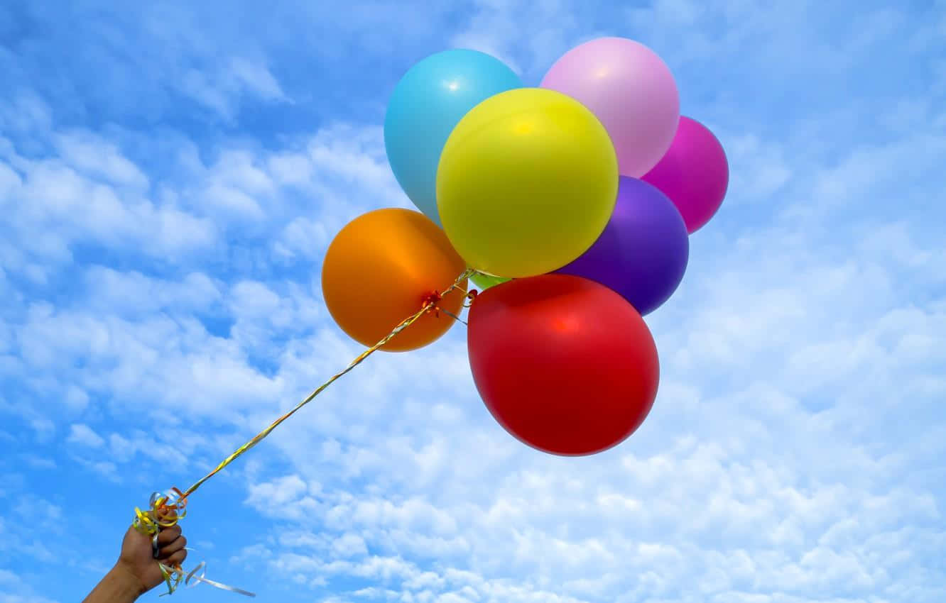 Balloon 桌布 壁紙 — 一隻手拿著一束鮮豔的彩虹氣球，背景是明亮的藍天和蓬鬆的白雲。