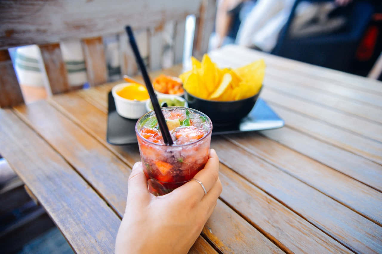 Food And Beverage 桌布 壁紙 — 一隻手拿著一杯充滿活力的紅色雞尾酒，配有冰塊和香草，放在一張風化的木桌上，旁邊有墨西哥玉米片和莎莎醬。