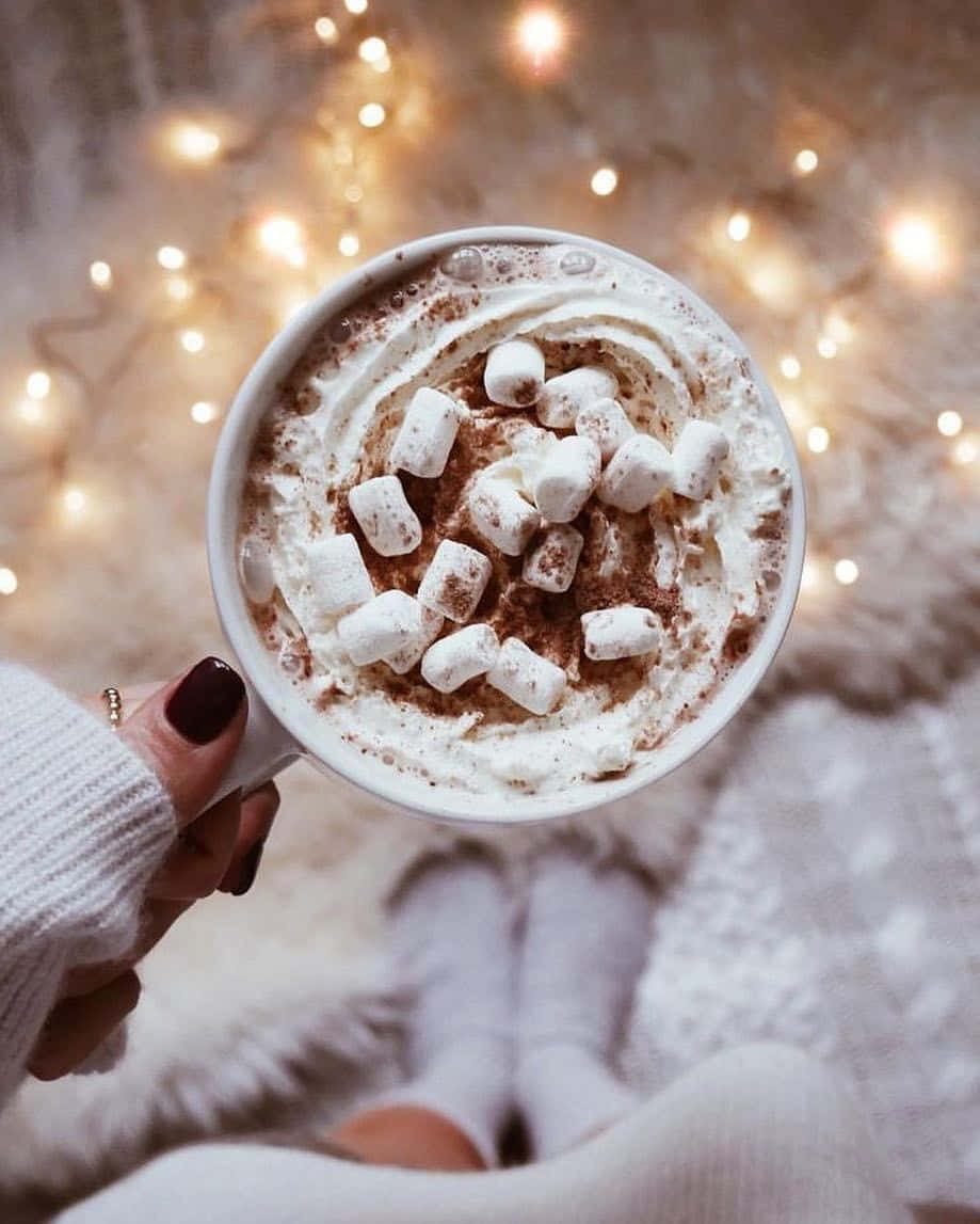 Hot Chocolate 桌布 壁紙 — 一隻手拿著一杯熱朱古力，上面有忌廉和迷你棉花糖，背景是溫馨的閃爍小燈。