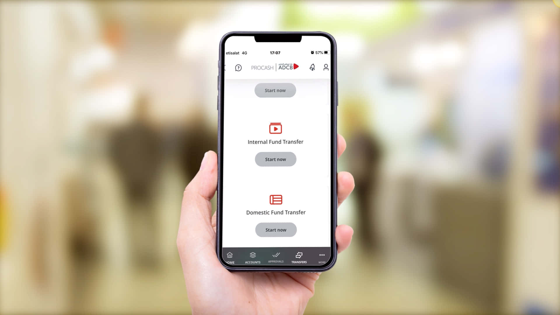 Mobile Banking 桌布 壁紙 — 一隻手拿著一部智能手機，顯示一個帶有轉賬選項的銀行應用程式，背景是模糊的室內環境。