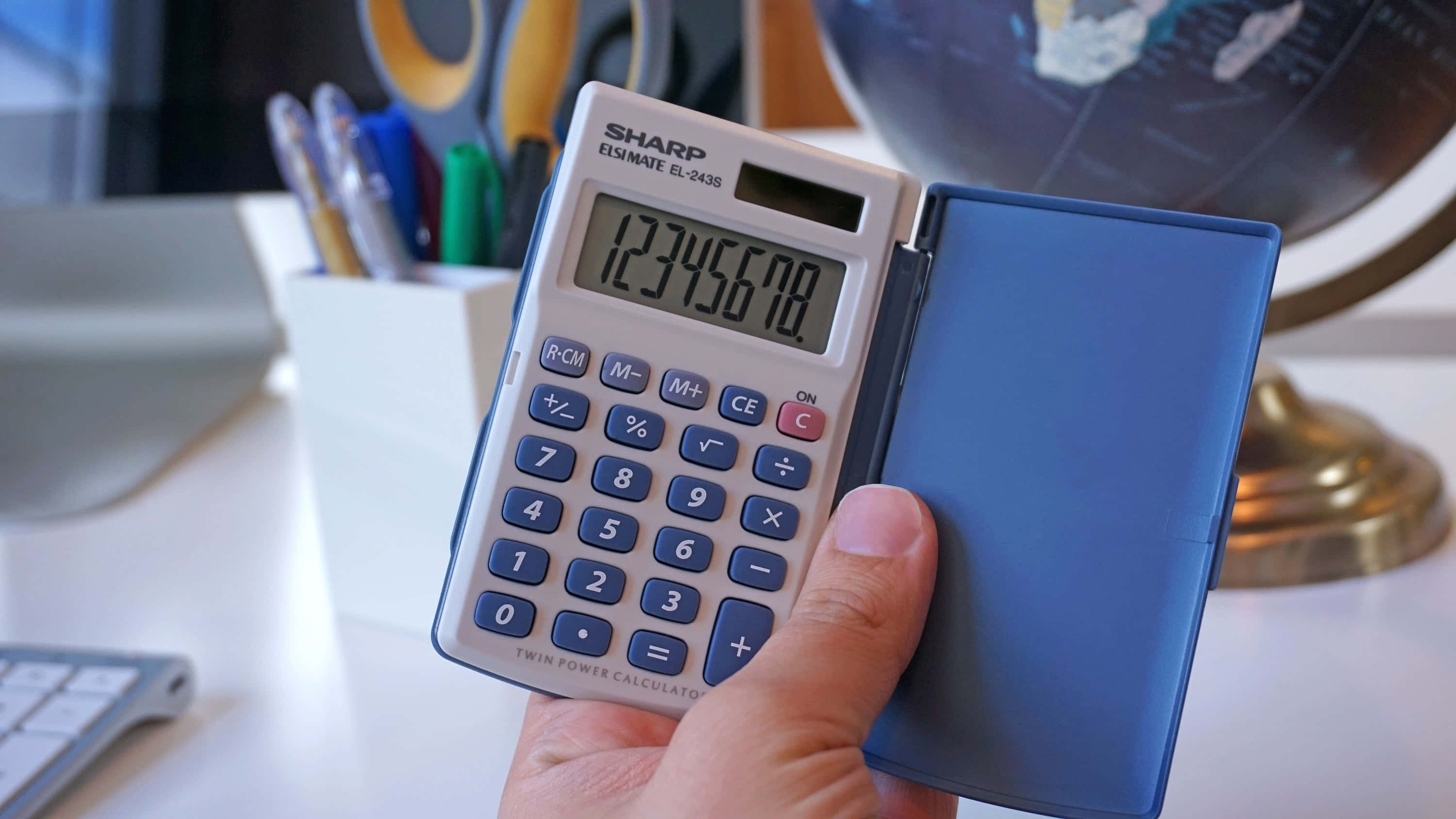 Calculator 桌布 壁紙 — 一隻手拿著一部顯示12345678的藍色Sharp計算機，旁邊有地球儀和筆，放在白色桌子上。