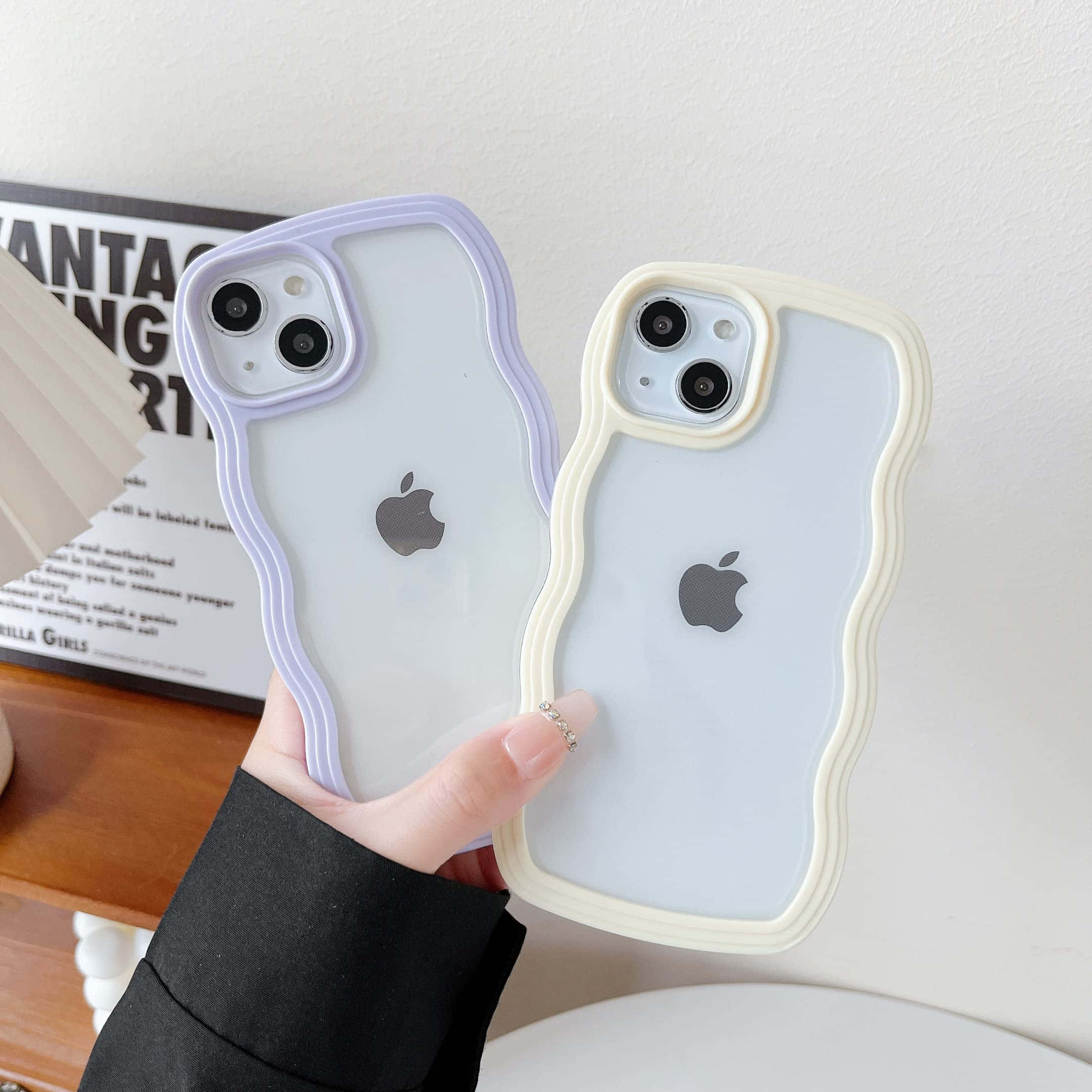 iPhone風格壁紙 桌布 壁紙 — 一隻手拿著兩個波浪邊半透明手機殼，一個薰衣草色一個奶油色，背景是白色牆壁，上面有海報和木架。