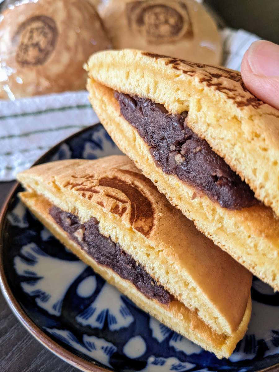 Japanese Sweets 桌布 壁紙 — 一隻手拿著半個金黃色煎餅，露出豐富的深紅色豆沙餡，放在藍白圖案碟上。