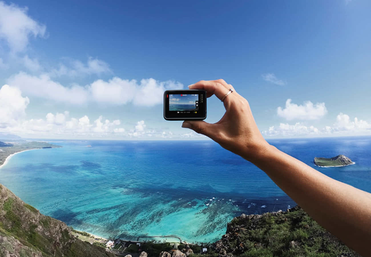 gopro 桌布 壁紙 — 一隻手拿著小型相機，捕捉令人驚嘆的海岸景色，包括碧綠的海水和遠處的火山島，在明亮的藍天下。