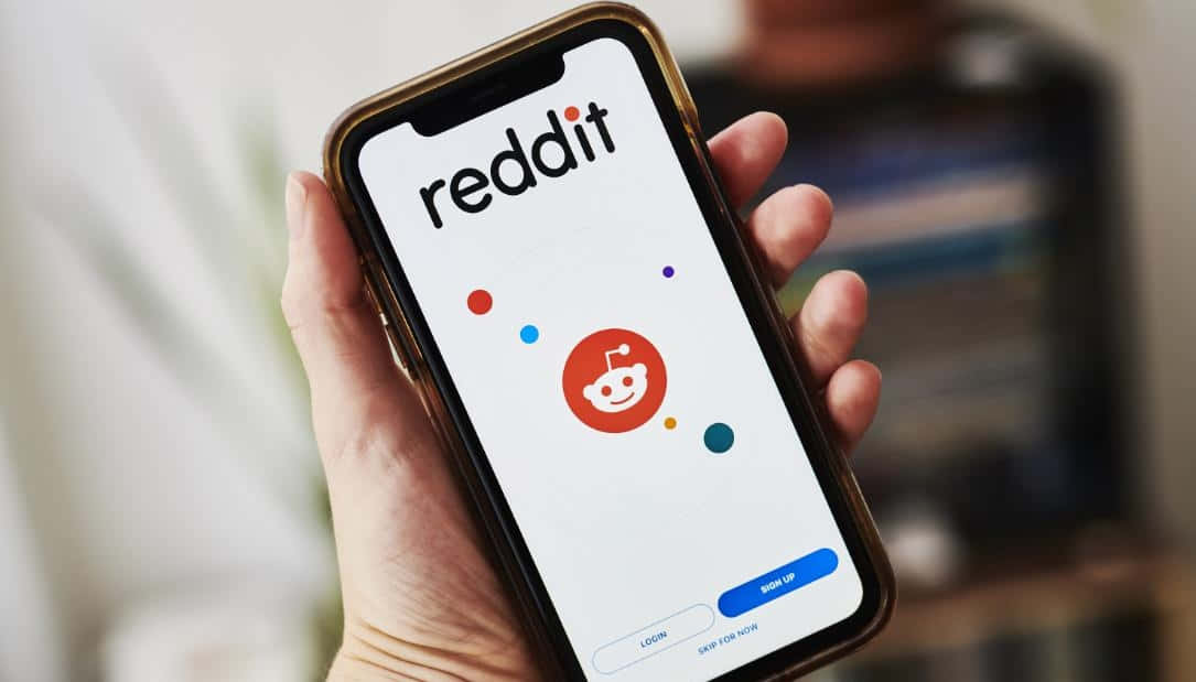 reddit 桌布 壁紙 — 一隻手拿著智能手機，顯示Reddit應用程式的登入畫面，帶有其標誌性的外星人吉祥物和彩色圓點。