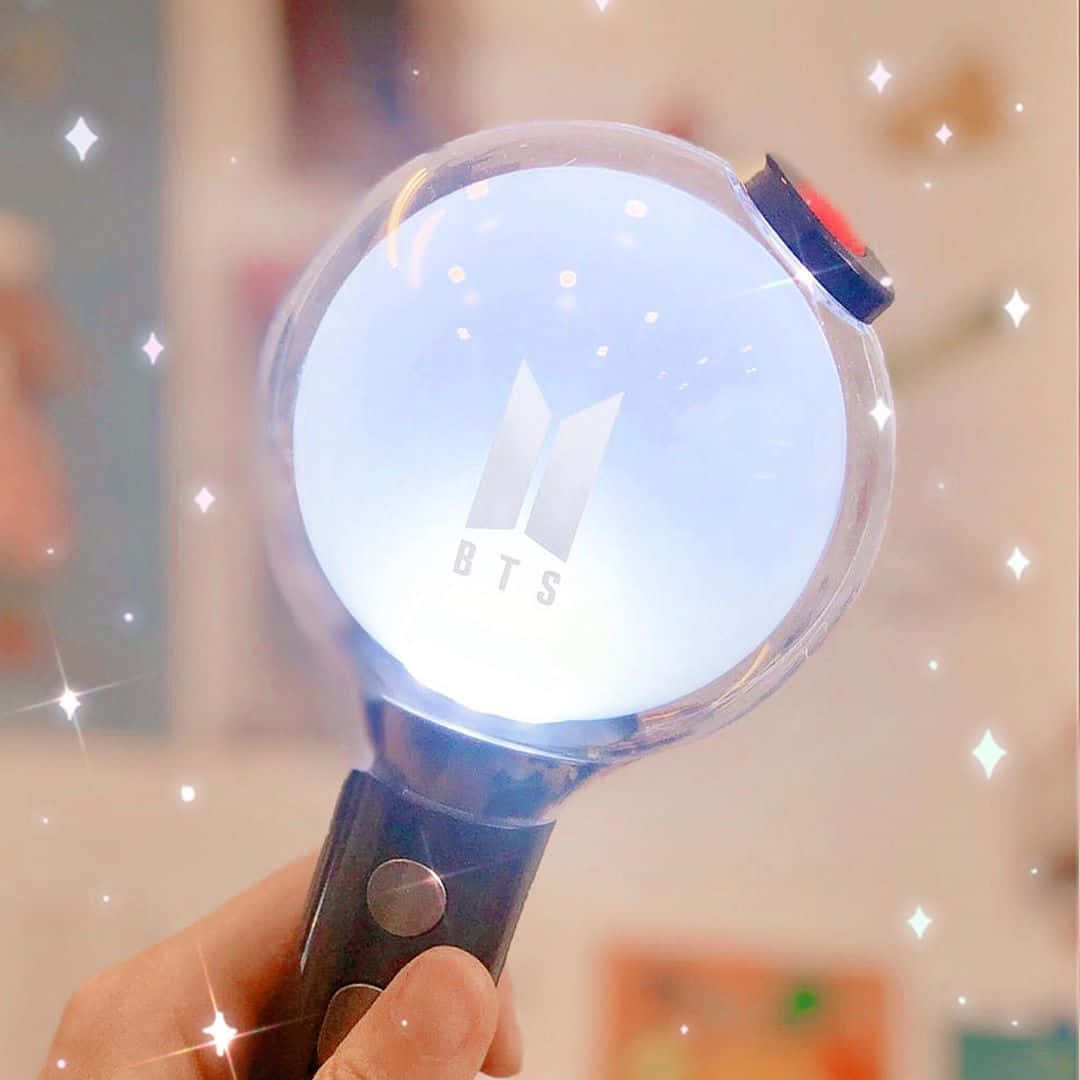 Bts Army Bomb 桌布 壁紙 — 一隻手拿著發光的BTS應援棒，帶有閃爍星星效果，背景柔和模糊。