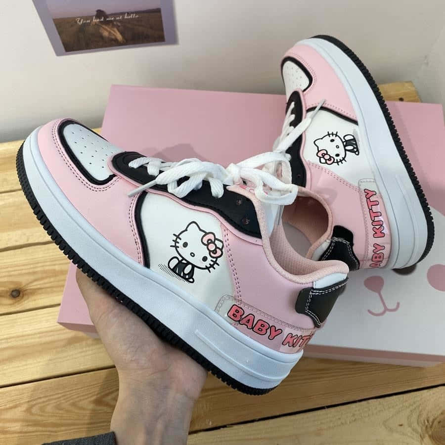 Kawaii Hello Kitty 桌布 壁紙 — 一隻手拿著粉紅、黑、白色的 Baby Kitty 運動鞋，上面有 Hello Kitty 圖案，背景係木質表面同粉紅色盒。