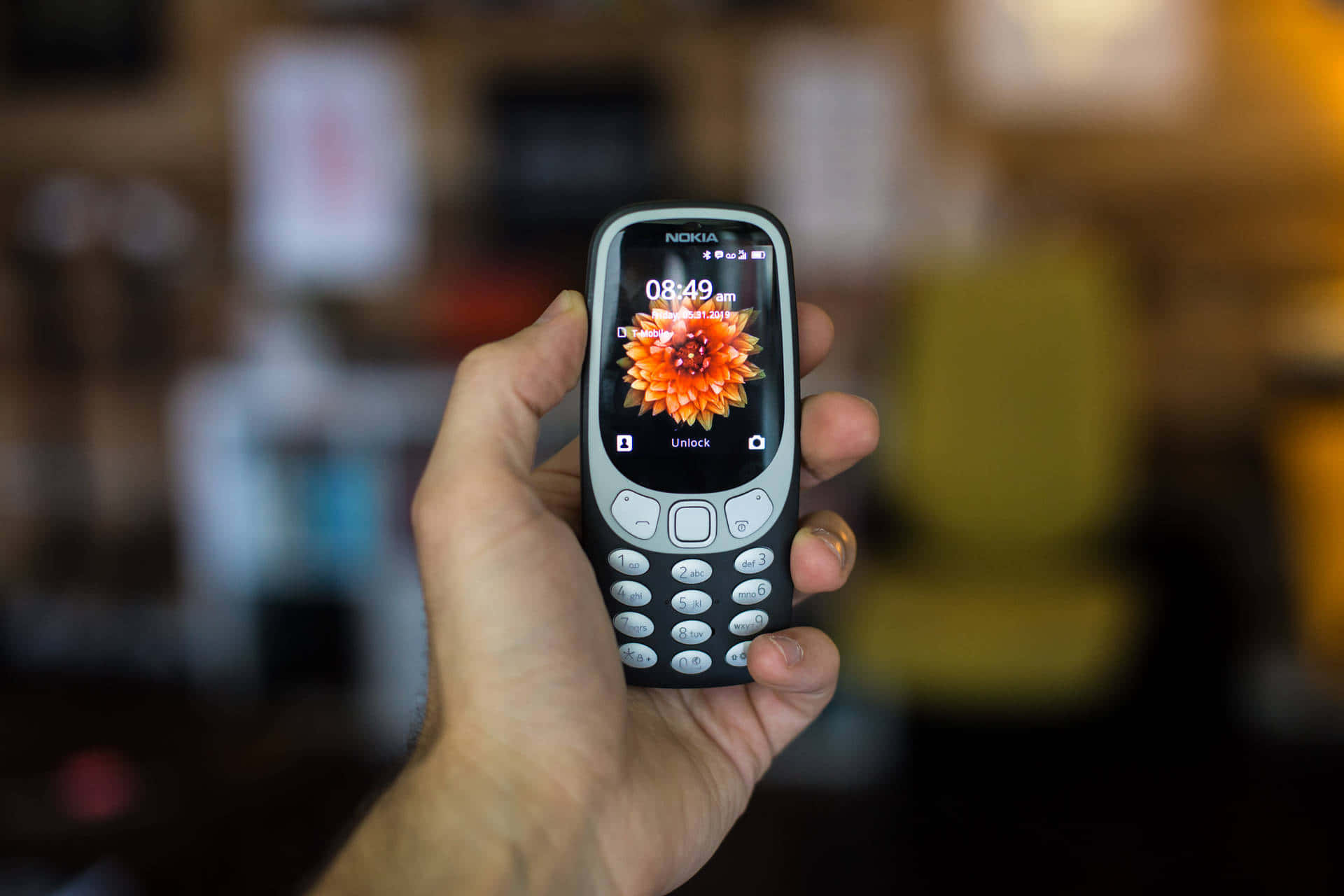 Nokia 桌布 壁紙 — 一隻手拿著經典的諾基亞手機，顯示充滿活力的橙色花朵桌布，背景是溫暖模糊的室內環境。