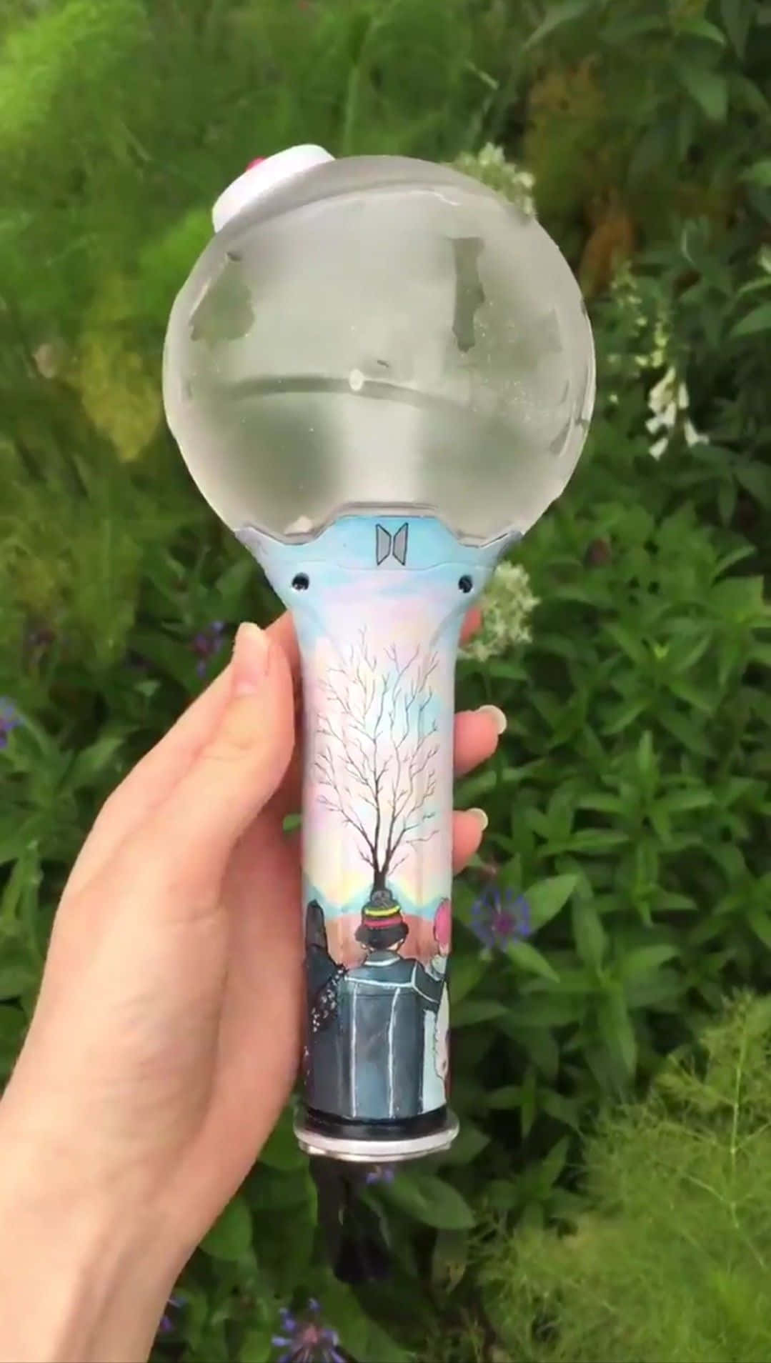 Bts Army Bomb 桌布 壁紙 — 一隻手拿著裝飾有樹木插圖的BTS應援棒，背景是茂密的綠色花園。