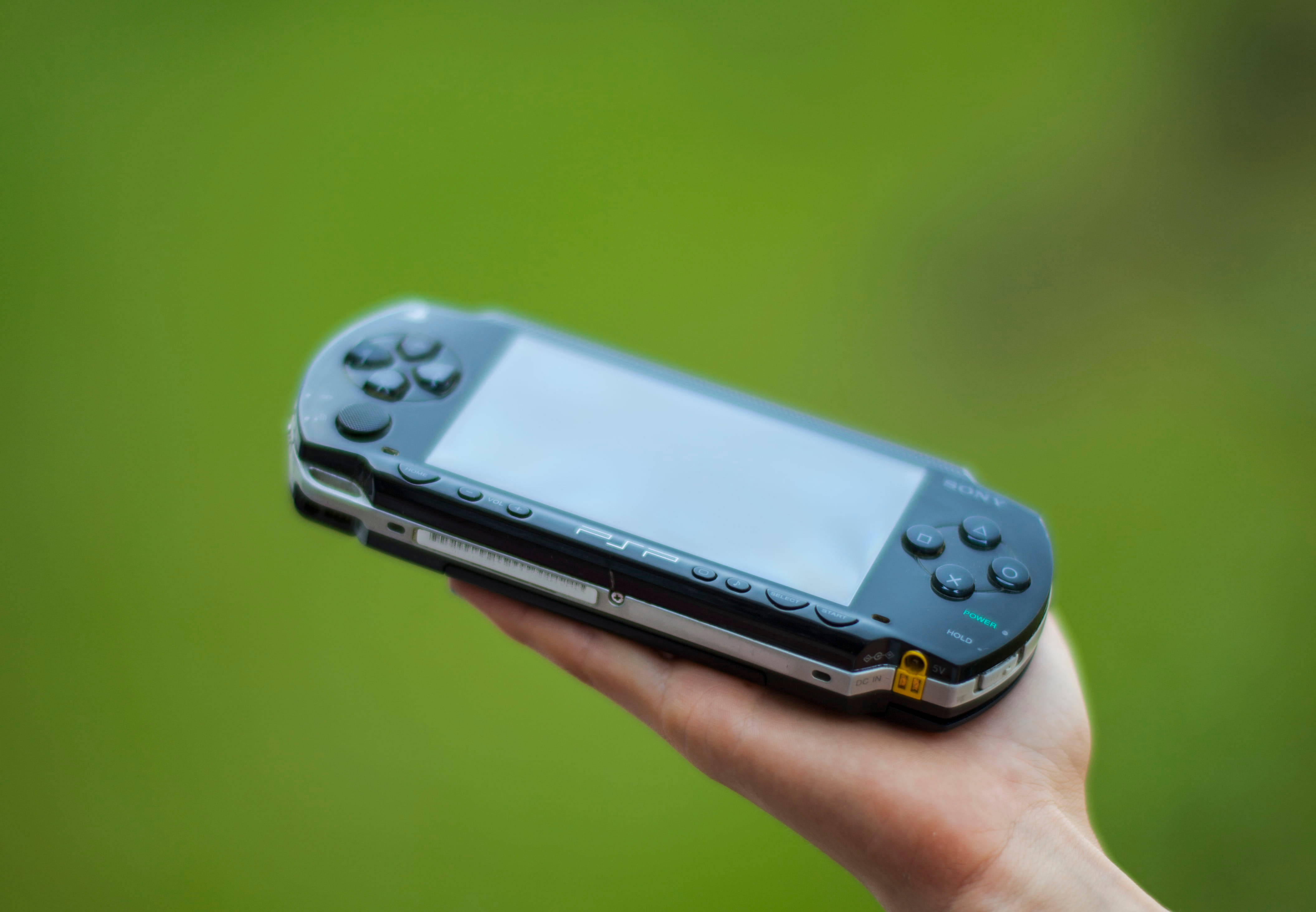 PlayStation 桌布 壁紙 — 一隻手拿著黑色Sony PSP手提遊戲機，背景是柔和的綠色模糊效果。