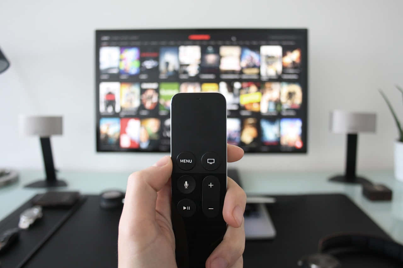 Home Entertainment 桌布 壁紙 — 一隻手拿著黑色遙控器，指向一個在現代客廳中顯示串流服務網格的電視。