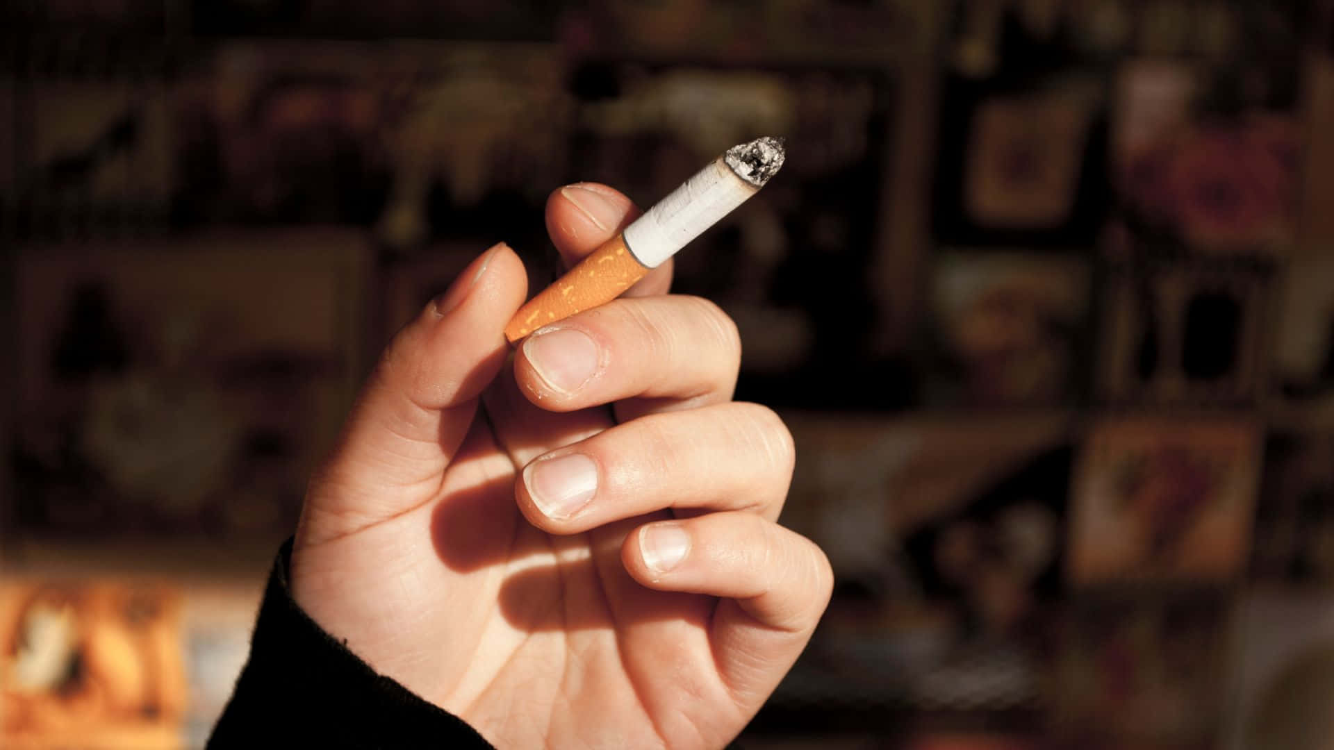 吸煙 桌布 壁紙 — 一隻手拿著點燃的香煙，背景是溫暖金色燈光下模糊的相框照片。