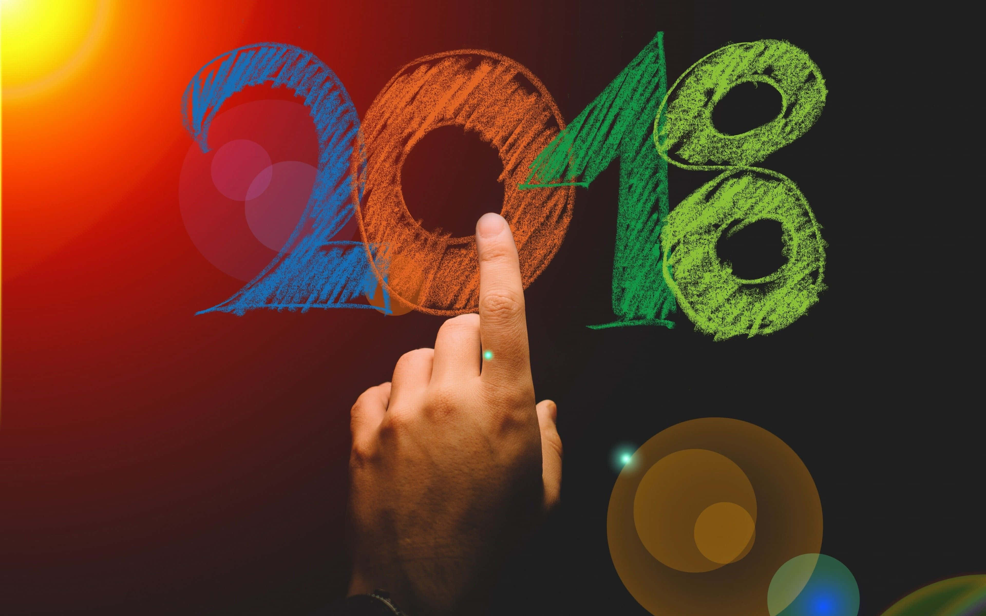 2018 桌布 壁紙 — 一隻手指向色彩繽紛的粉筆風格「2018」，背景是充滿活力的漸變色並帶有鏡頭光暈。