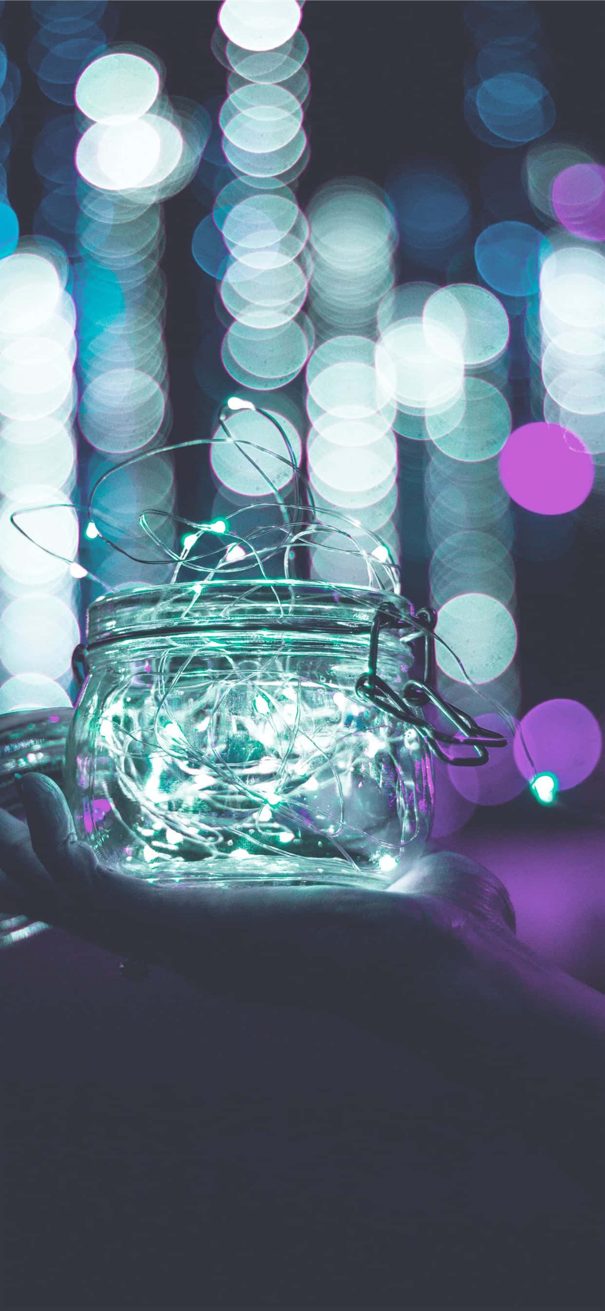 Spring Lights 桌布 壁紙 — 一隻手捧著一個裝滿發光仙女燈的玻璃罐，背景是紫色和藍色燈光構成的鮮豔散景。