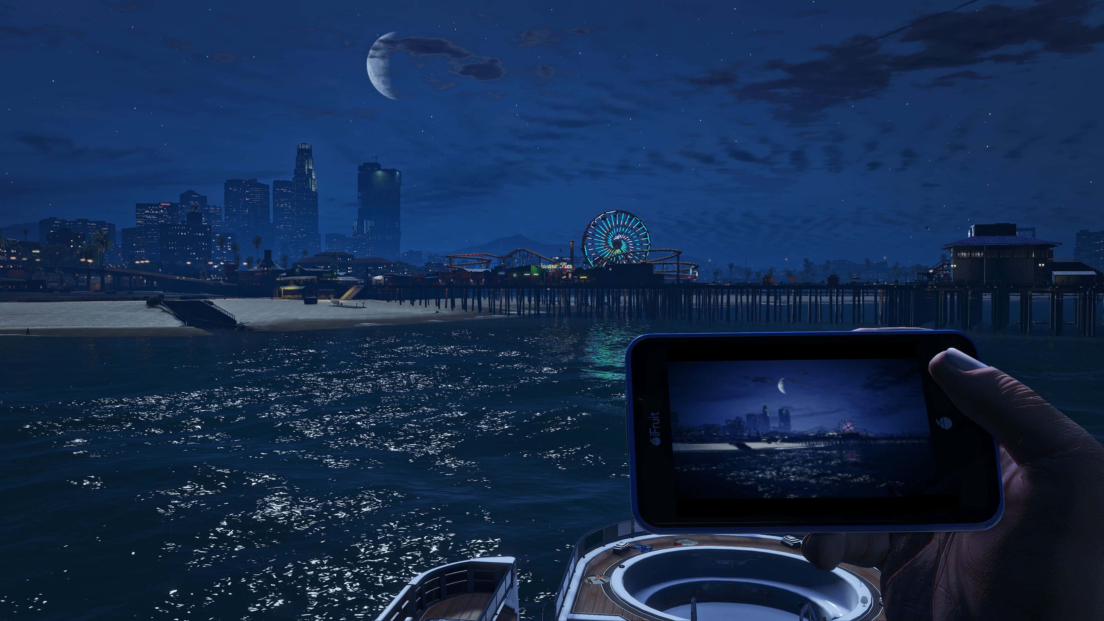 Gta V 4K 桌布 壁紙 — 一隻手握住手機，捕捉月光下閃爍摩天輪和波光粼粼水面的海岸城市夜景。