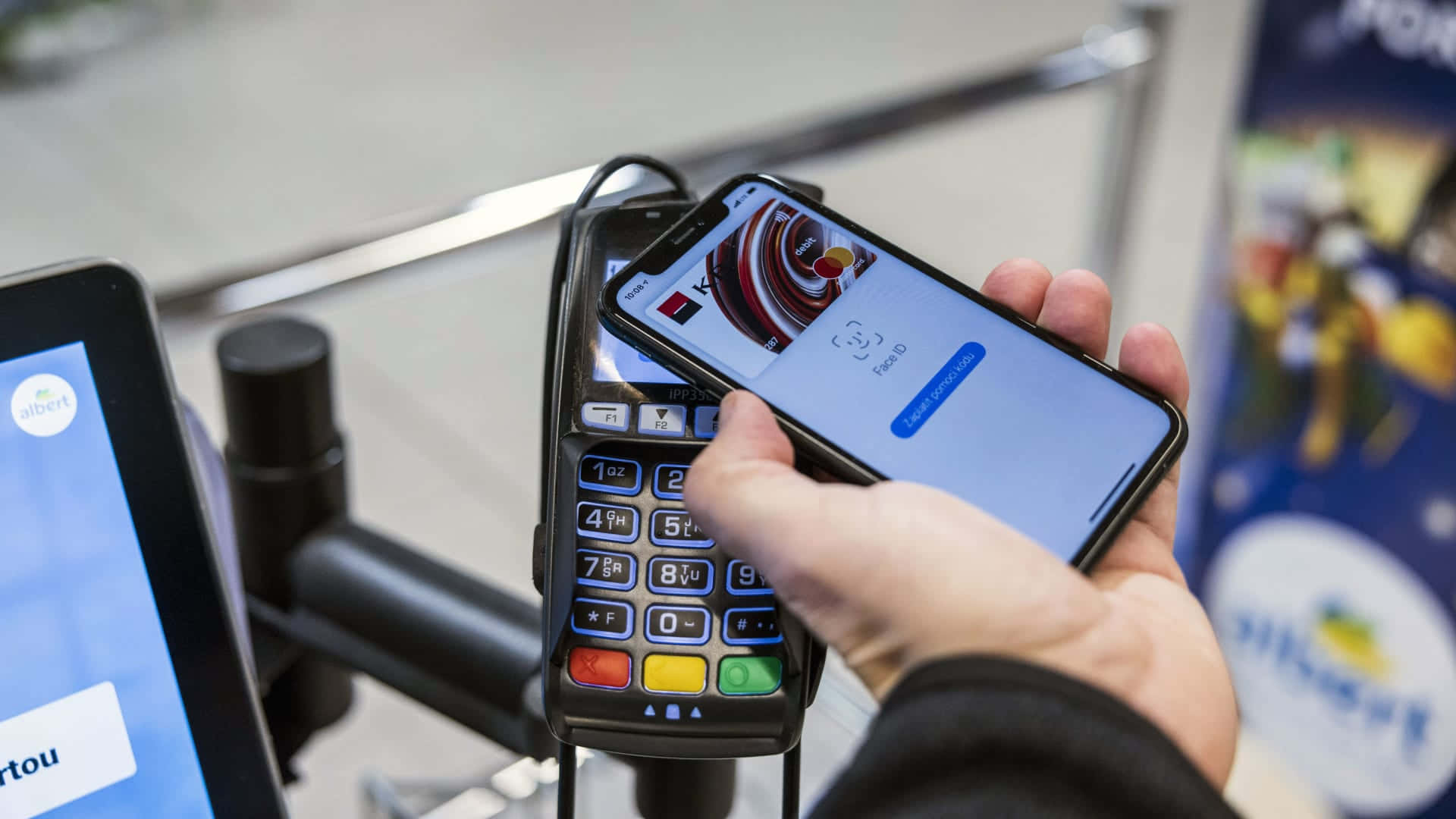 iPhone Apple Pay Bloomberg 壁紙 — 一隻手握住智能手機，在現代零售環境中輕觸非接觸式卡於支付終端上。
