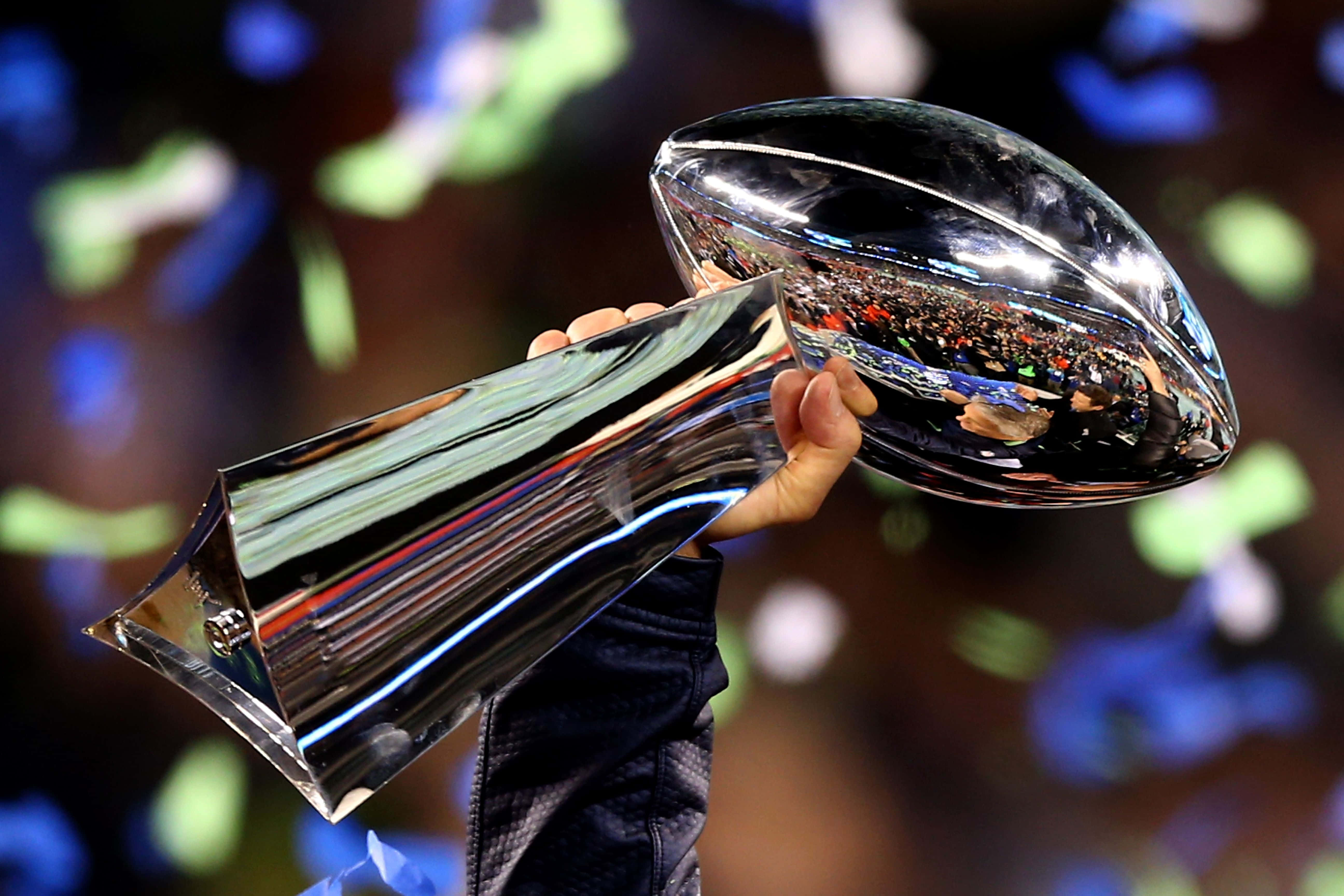 Super Bowl 桌布 壁紙 — 一隻手握住閃閃發光的銀色超級碗獎盃，五彩紙屑在充滿歡呼球迷的體育館中飄落。