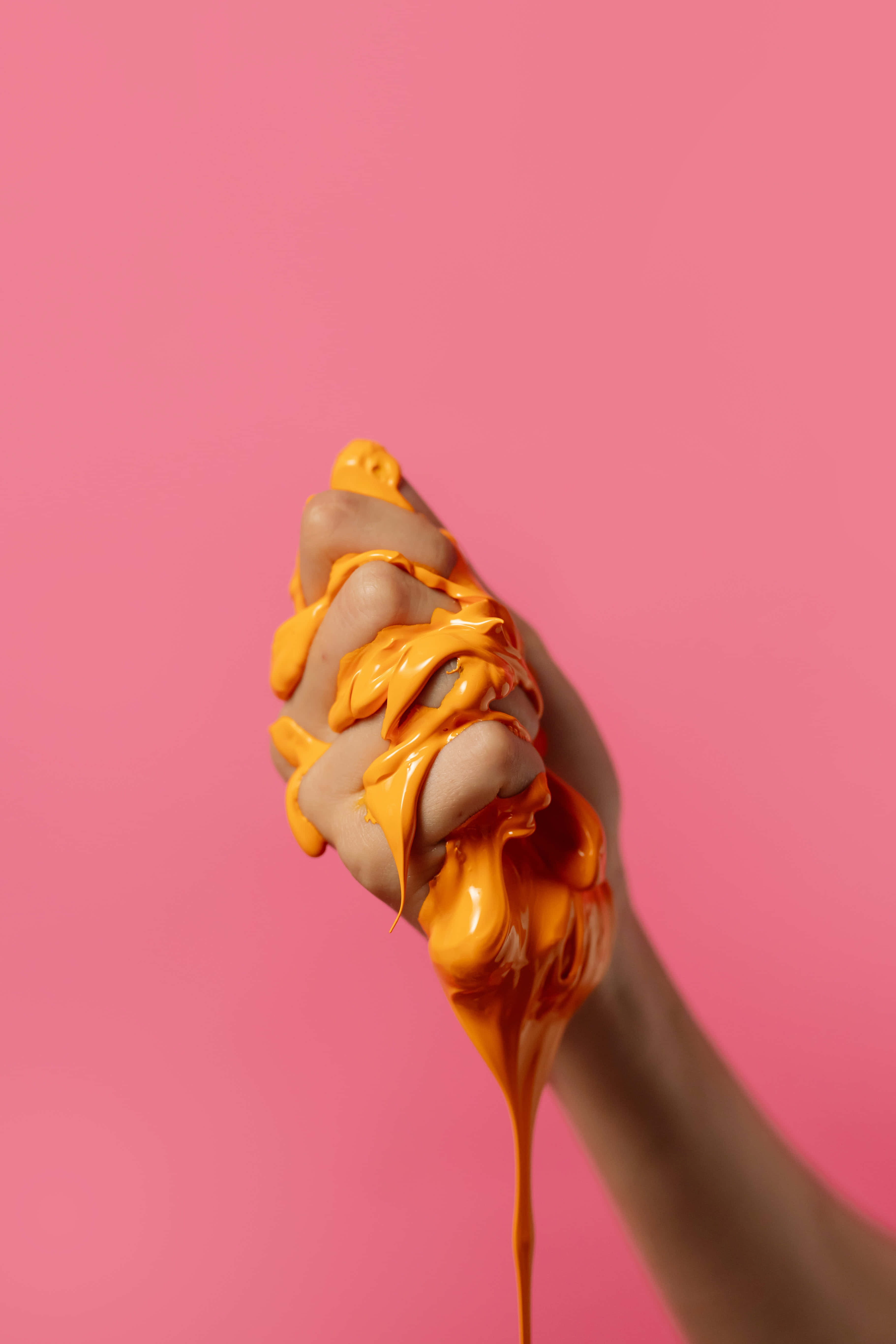 slime 桌布 壁紙 — 一隻手握成拳頭，上面覆蓋著滴落的橙色油漆，背景為純粉色。