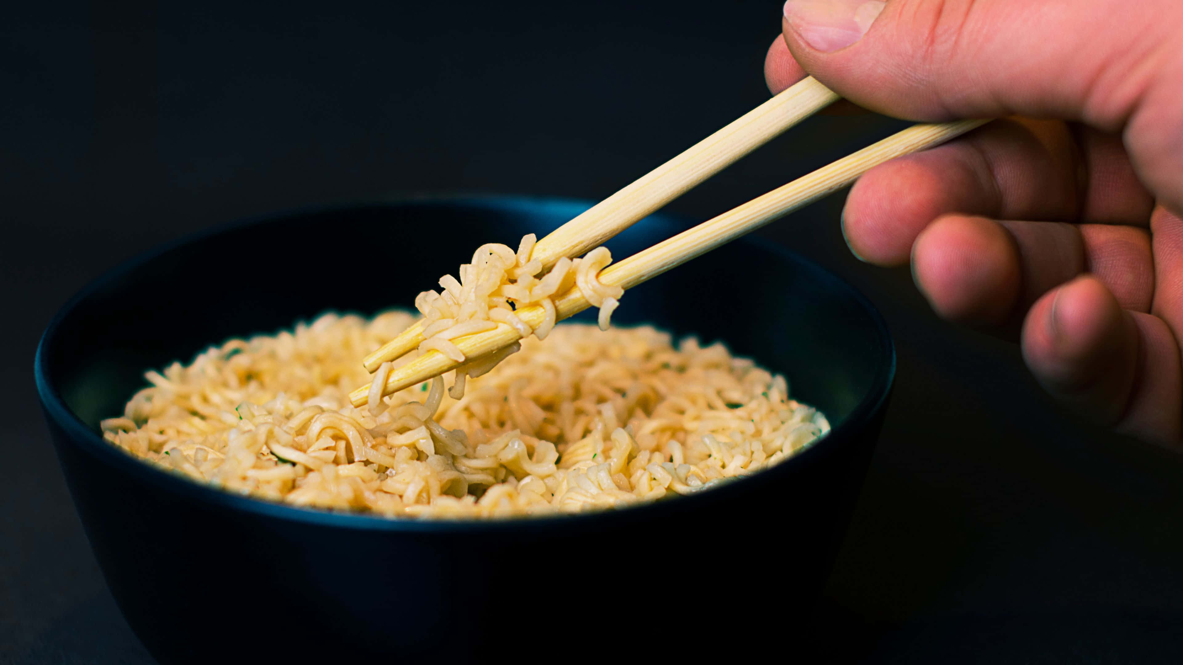 Noodles 桌布 壁紙 — 一隻手用筷子從黑色背景的深色碗中夾起冒煙的即食麵。