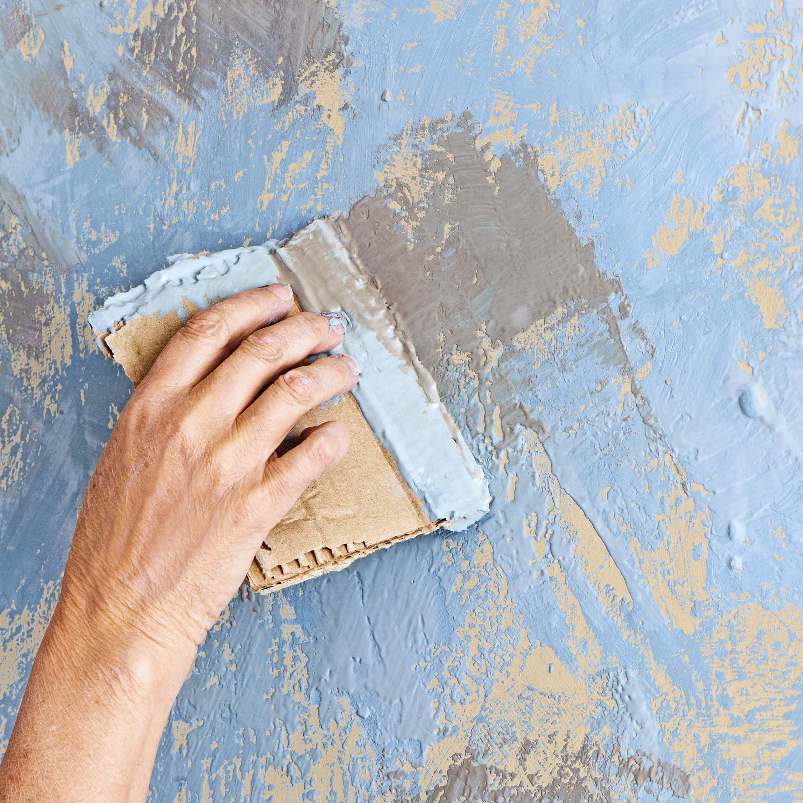 Paint Texture 桌布 壁紙 — 一隻手用紙板刮刀將有紋理的藍色油漆塗在風化且露出米色斑塊的牆上。
