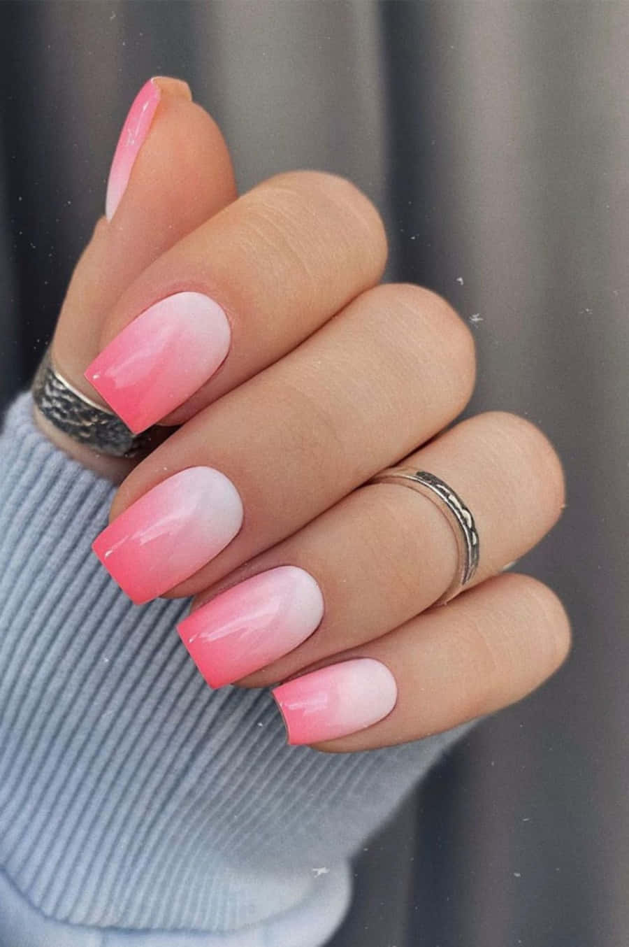 Pink Nail Art 桌布 壁紙 — 一隻手的特寫，帶有柔和粉紅漸變指甲和銀戒指，背景為柔和灰色。
