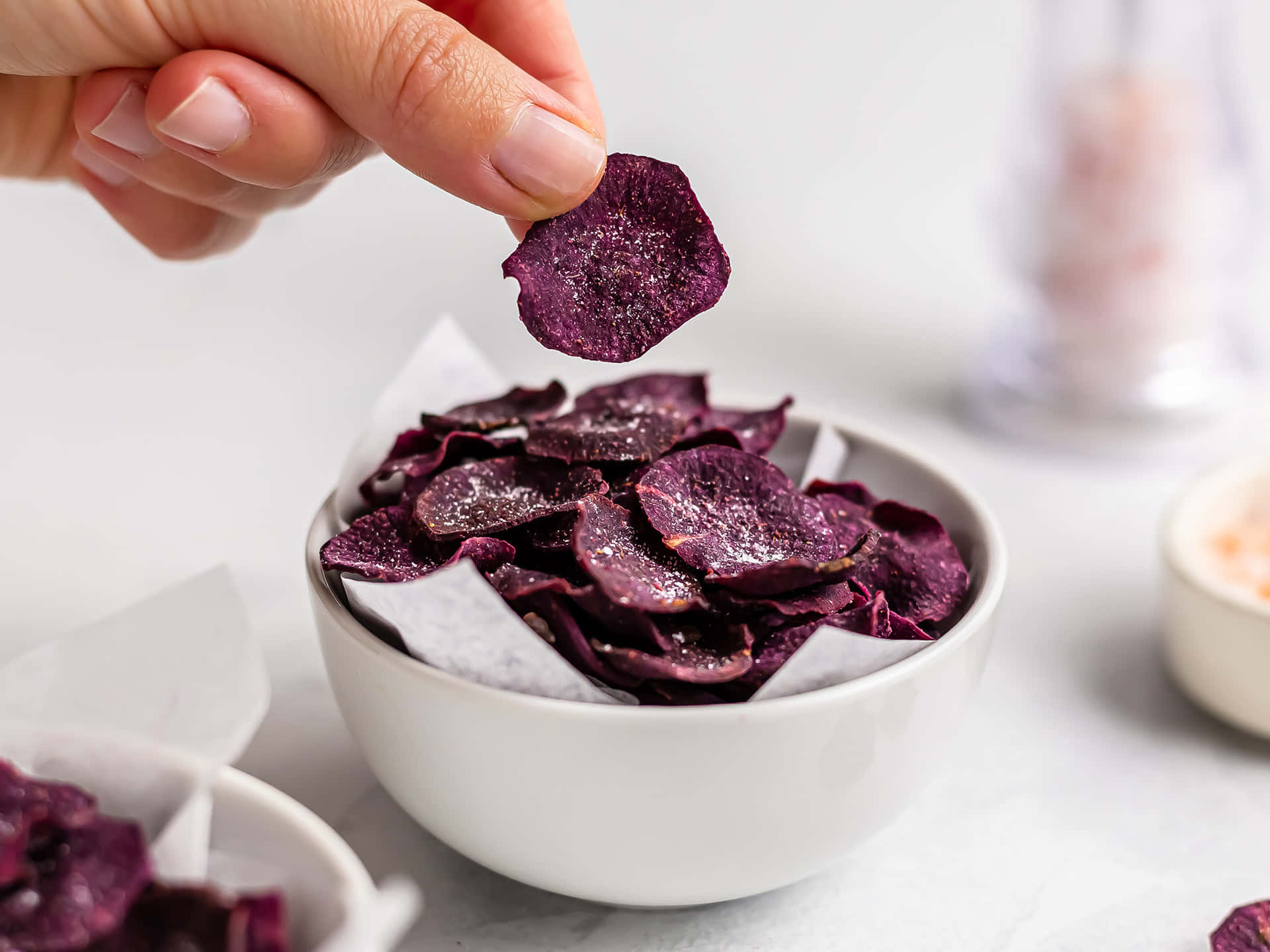 Purple Sweet Potato 桌布 壁紙 — 一隻手輕輕地從鋪有烘焙紙的白色碗中拿起一塊閃閃發光的紫色番薯片。