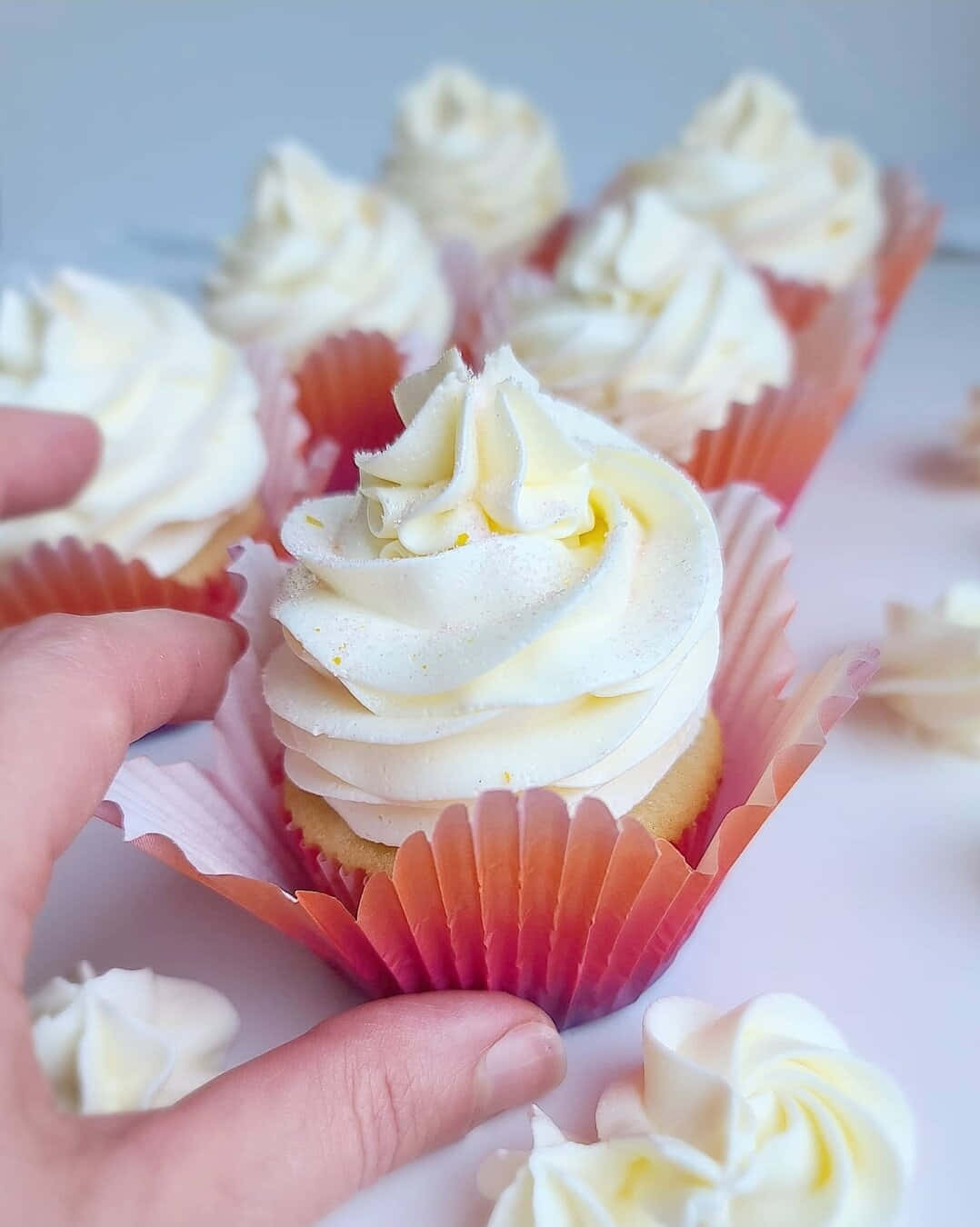 Buttercream 桌布 壁紙 — 一隻手輕輕拿著一個帶有奶油白色糖霜和橙色紙杯的杯子蛋糕，背景是柔和的粉彩色。