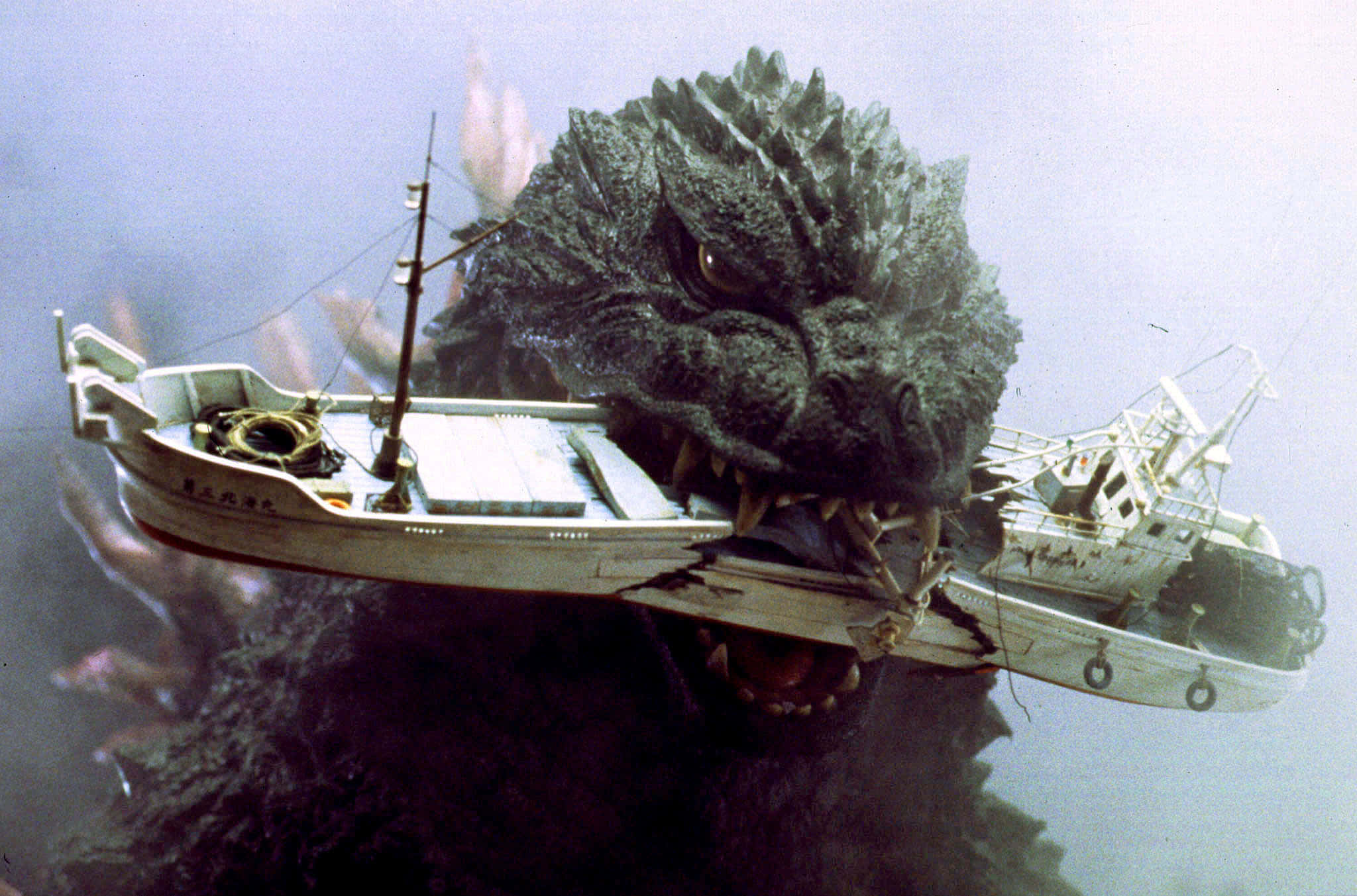 Godzilla 2000 桌布 壁紙 — 一隻擁有尖刺灰色皮膚的怪獸哥斯拉在渾濁的海水中咬住一艘小漁船。