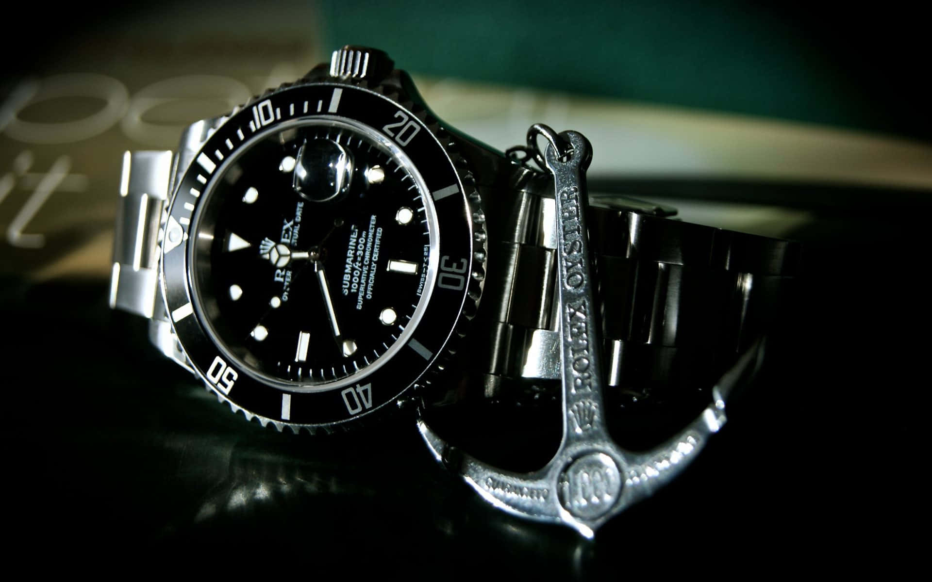 Rolex 桌布 壁紙 — 一隻時尚的黑色勞力士Submariner手錶在戲劇性燈光下，於深色反光表面旁與銀色錨形鑰匙扣一同閃耀。