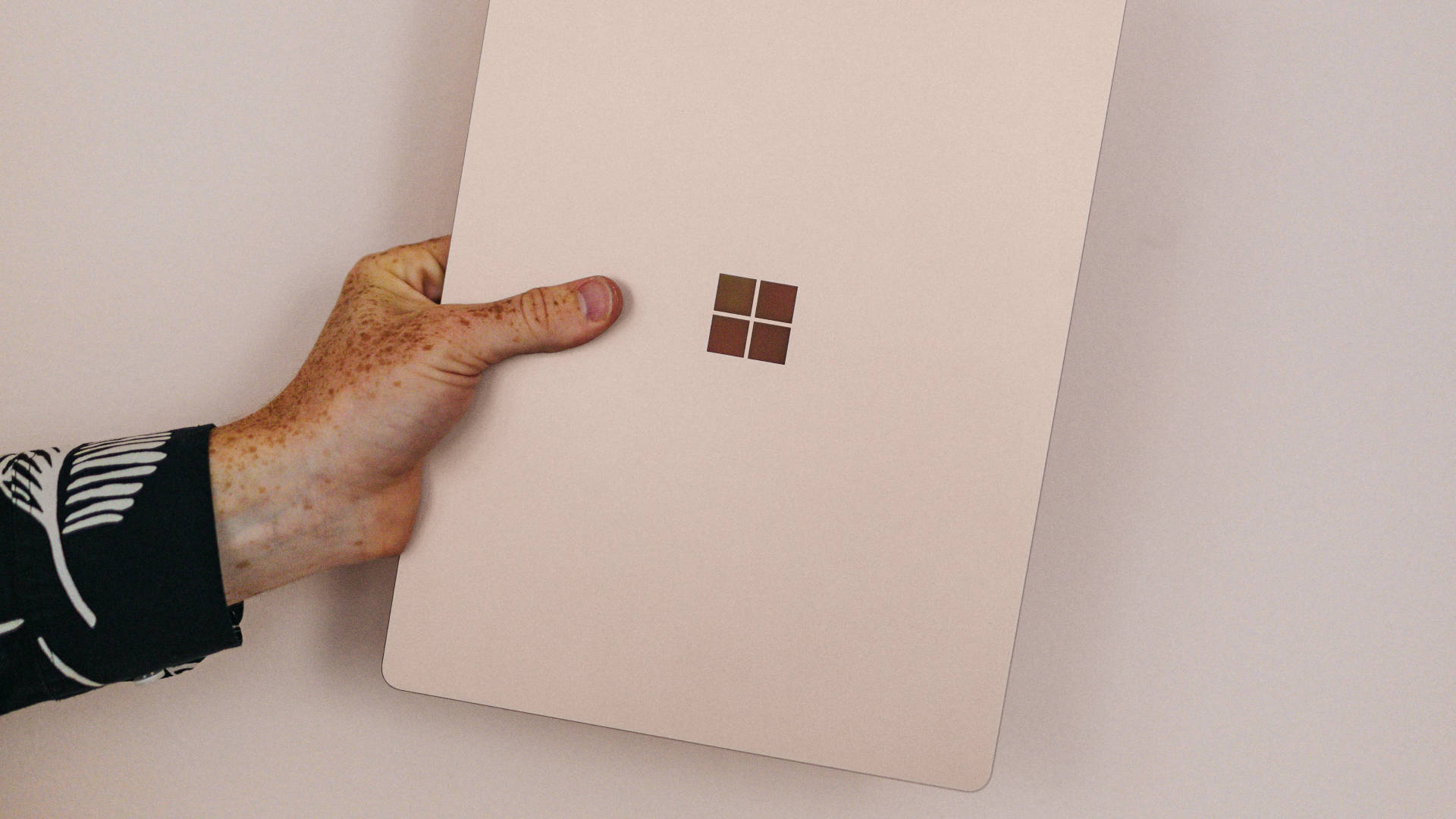 microsoft 桌布 壁紙 — 一隻有雀斑的手拿著淺粉紅色的Microsoft Surface手提電腦，靠著素色牆壁。