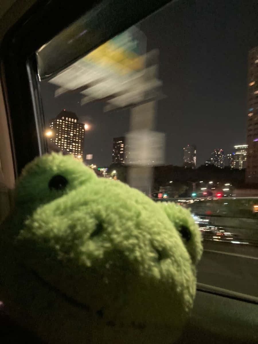 Aesthetic Frog 桌布 壁紙 — 一隻毛茸茸的綠色毛絨青蛙從火車窗戶探出頭來，夜晚模糊的城市燈光呼嘯而過。
