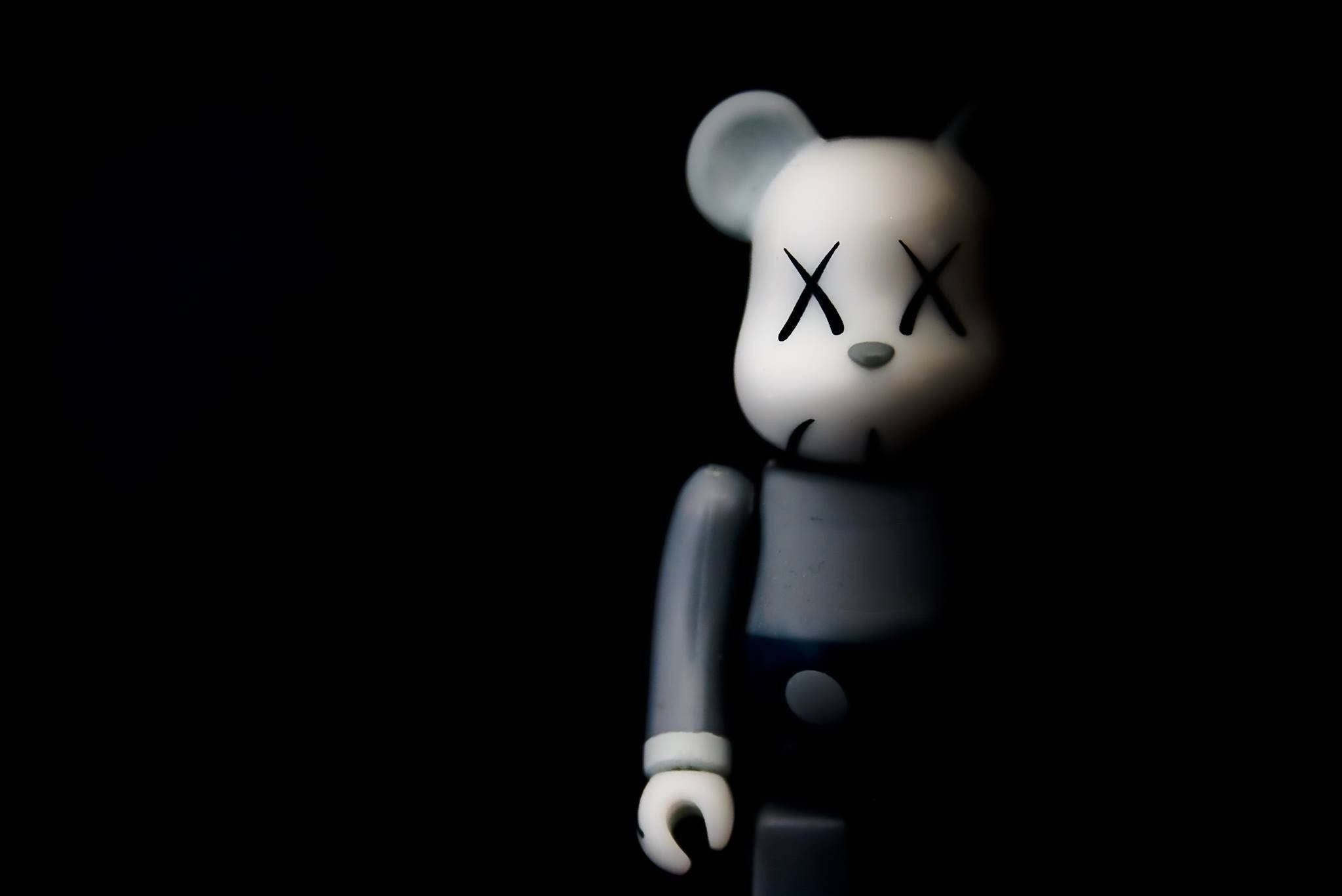 Bearbrick KAWS 46 壁紙 — 一隻白色KAWS熊公仔，帶有X形眼睛，在昏暗光線下站在黑色背景前。