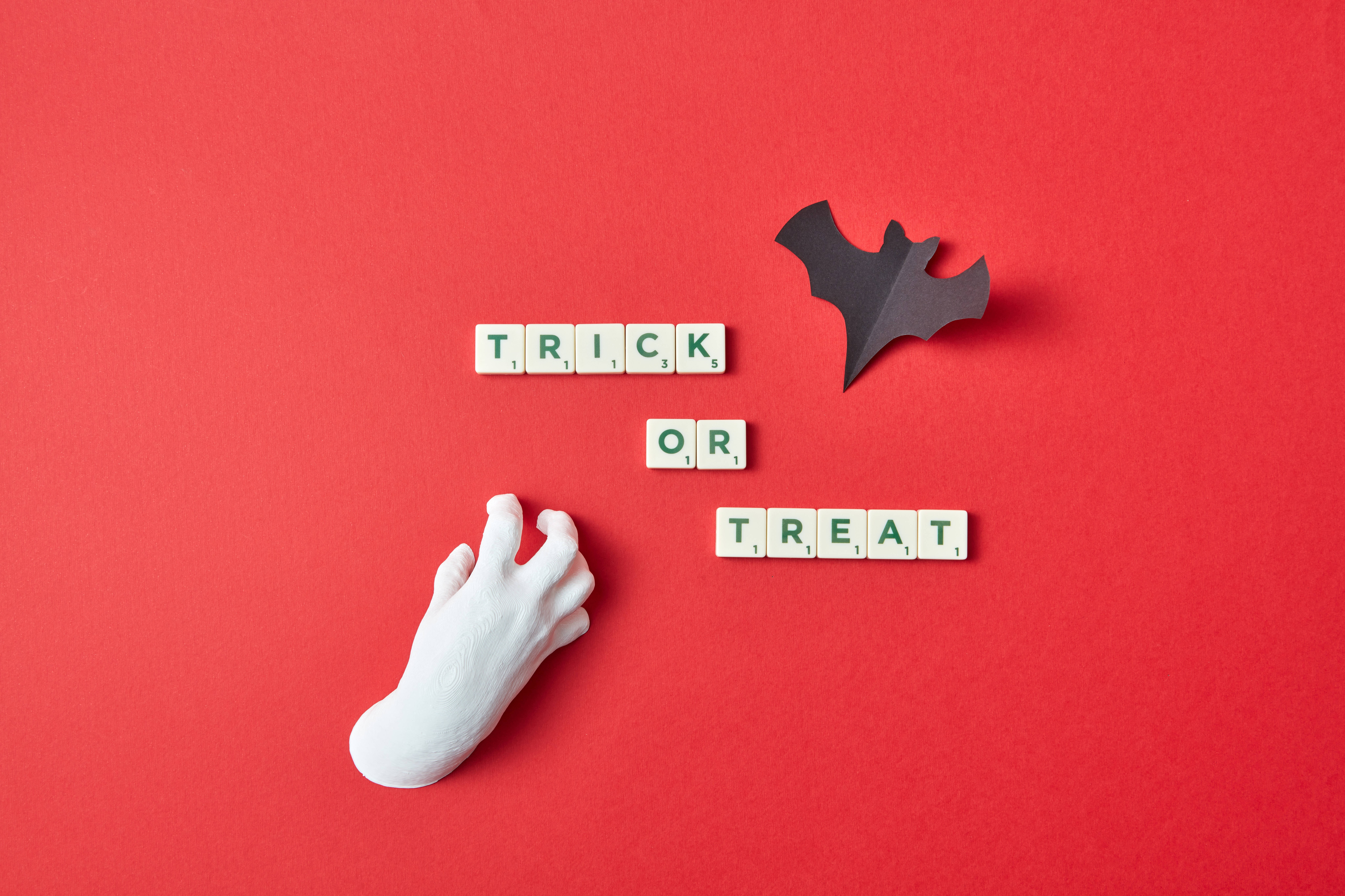 萬聖節 桌布 壁紙 — 一隻白色塑膠手伸向拼出 trick or treat 的 Scrabble 字母磚，背景為大膽的紅色，並有一隻黑色紙蝙蝠。