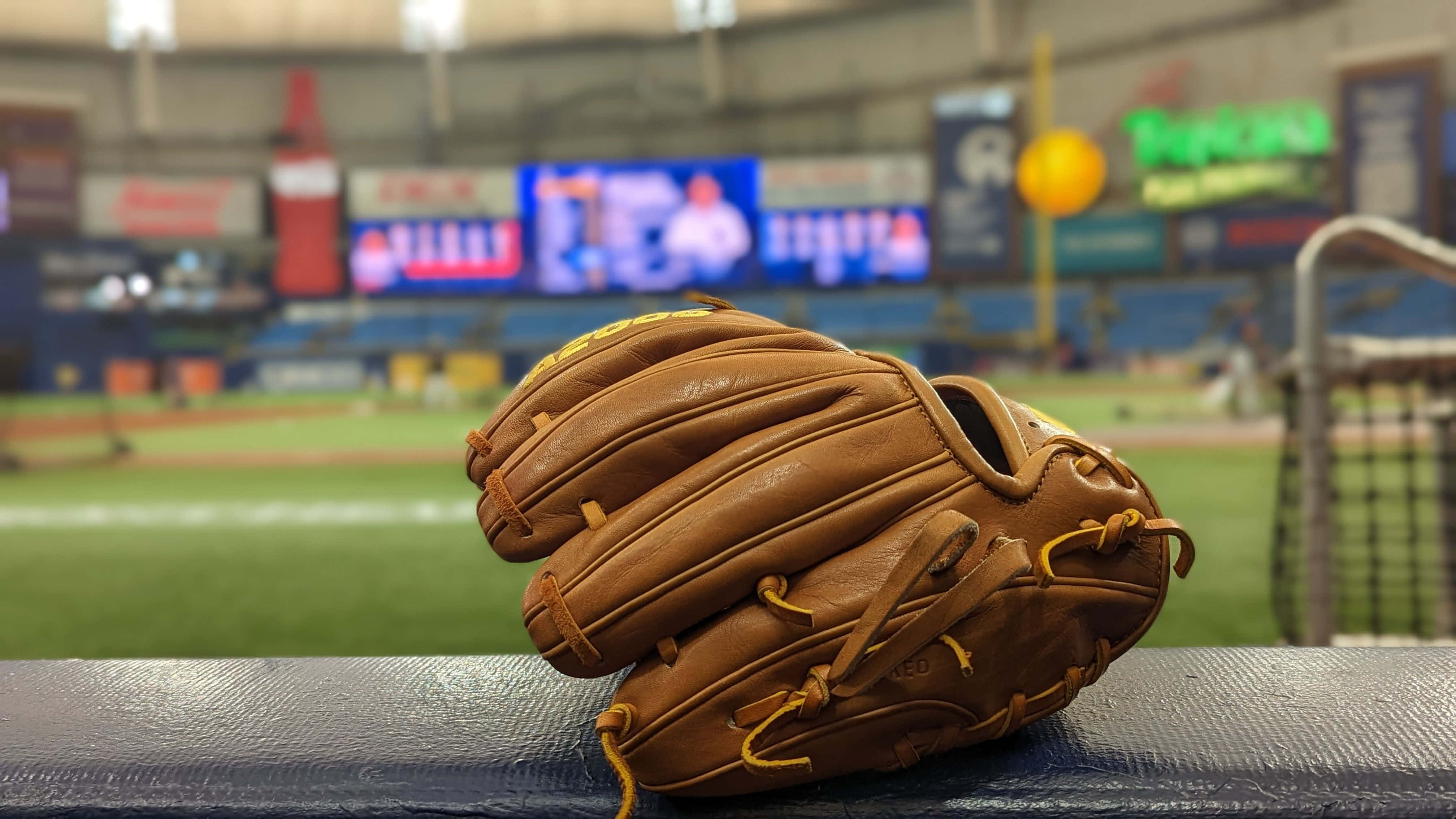 Baseball Gloves 桌布 壁紙 — 一隻磨損的棕色棒球手套放在球員休息區的長椅上，背景是模糊的球場和記分板。