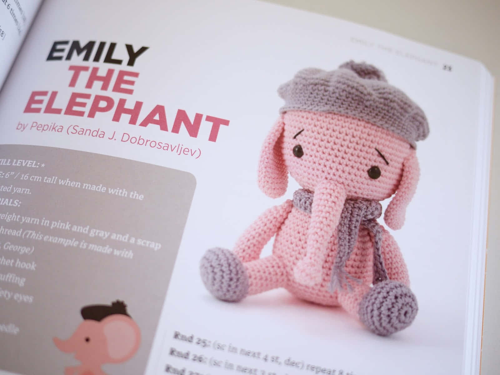 Emily Elephant 桌布 壁紙 — 一隻粉紅色鉤針大象戴着灰色帽子和圍巾，坐在一本名為《Emily the Elephant》的手工書頁旁邊。