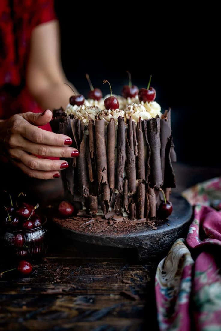 Black Forest Cake 桌布 壁紙 — 一隻紅指甲的手在深色鄉村背景下輕輕拿著一塊裝飾有櫻桃和奶油的頹廢朱古力蛋糕。