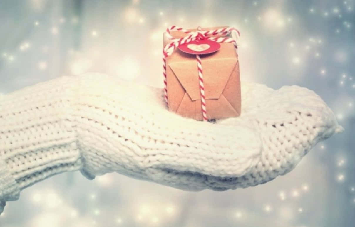 Mittens 桌布 壁紙 — 一隻舒適的白色編織手輕輕拿著一個用紅白繩子綁著的小棕色禮物盒，背景是閃閃發光的冬日場景。