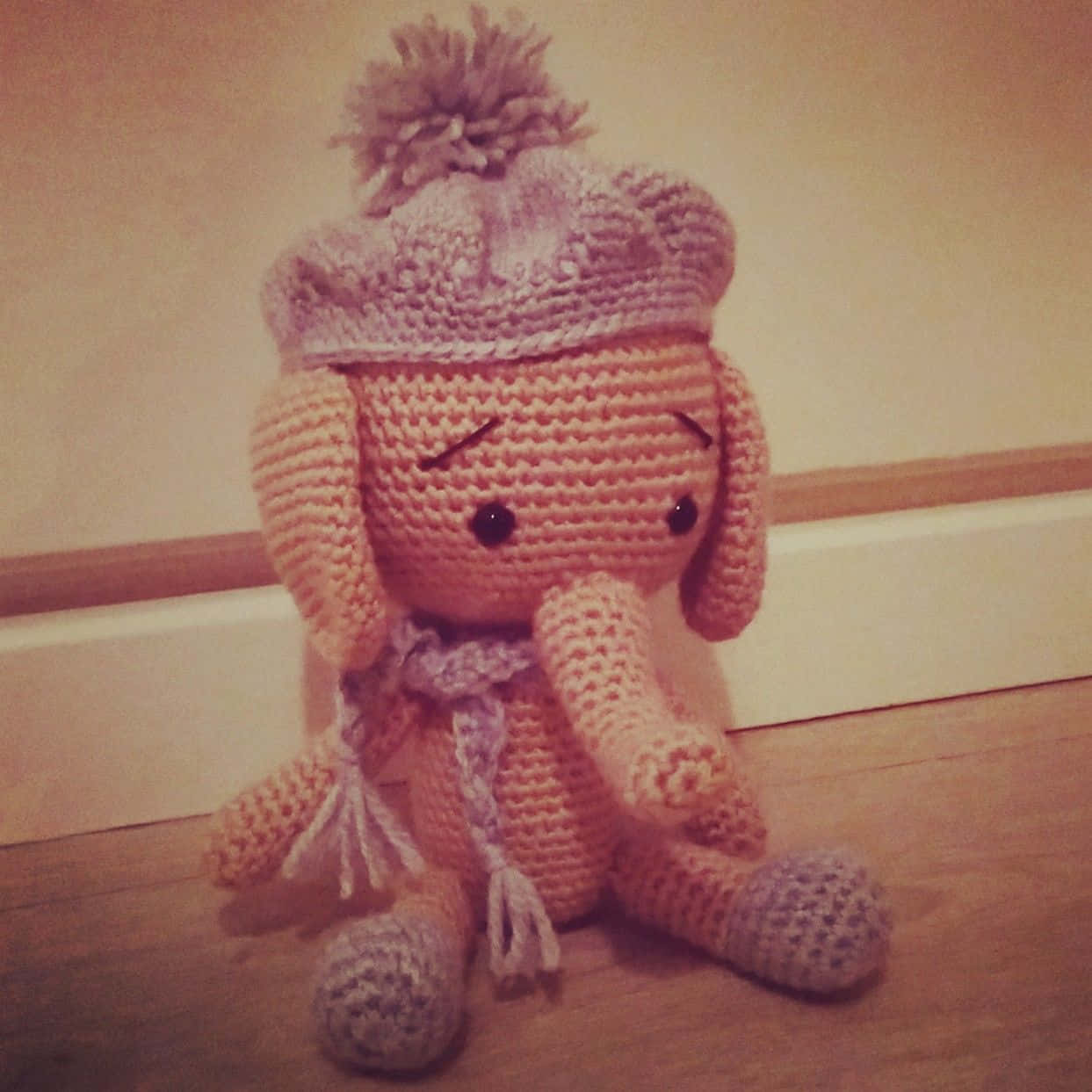 Emily Elephant 桌布 壁紙 — 一隻舒適的鉤針編織大象毛絨玩具，戴著帶有絨球的針織帽，坐在踢腳線附近的木地板上。