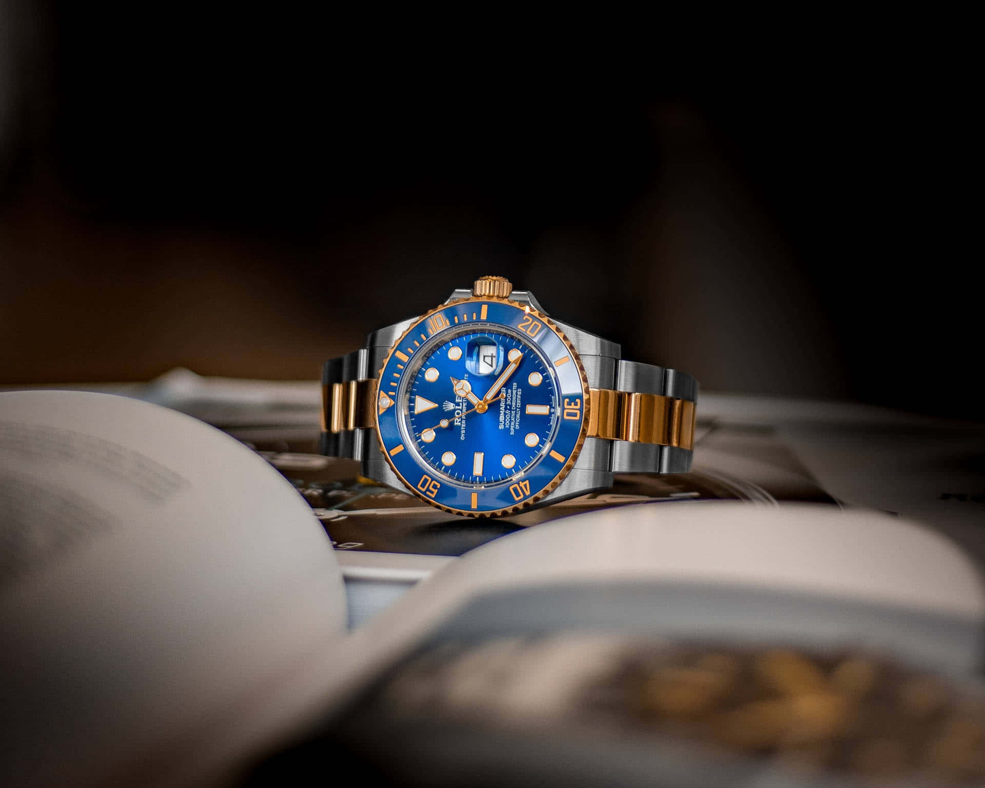 Rolex 桌布 壁紙 — 一隻豪華勞力士手錶，帶有藍色錶盤和金色點綴，優雅地放在一本打開的書上，環境昏暗。