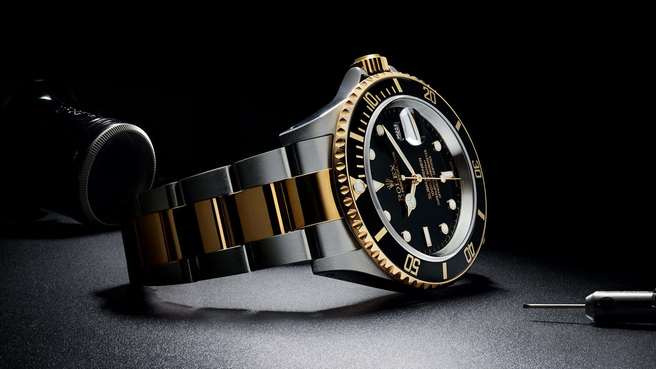 Rolex 桌布 壁紙 — 一隻豪華的金色和鋼製勞力士手錶戲劇性地放在黑暗的表面上，附近有工具，在聚焦的燈光下。