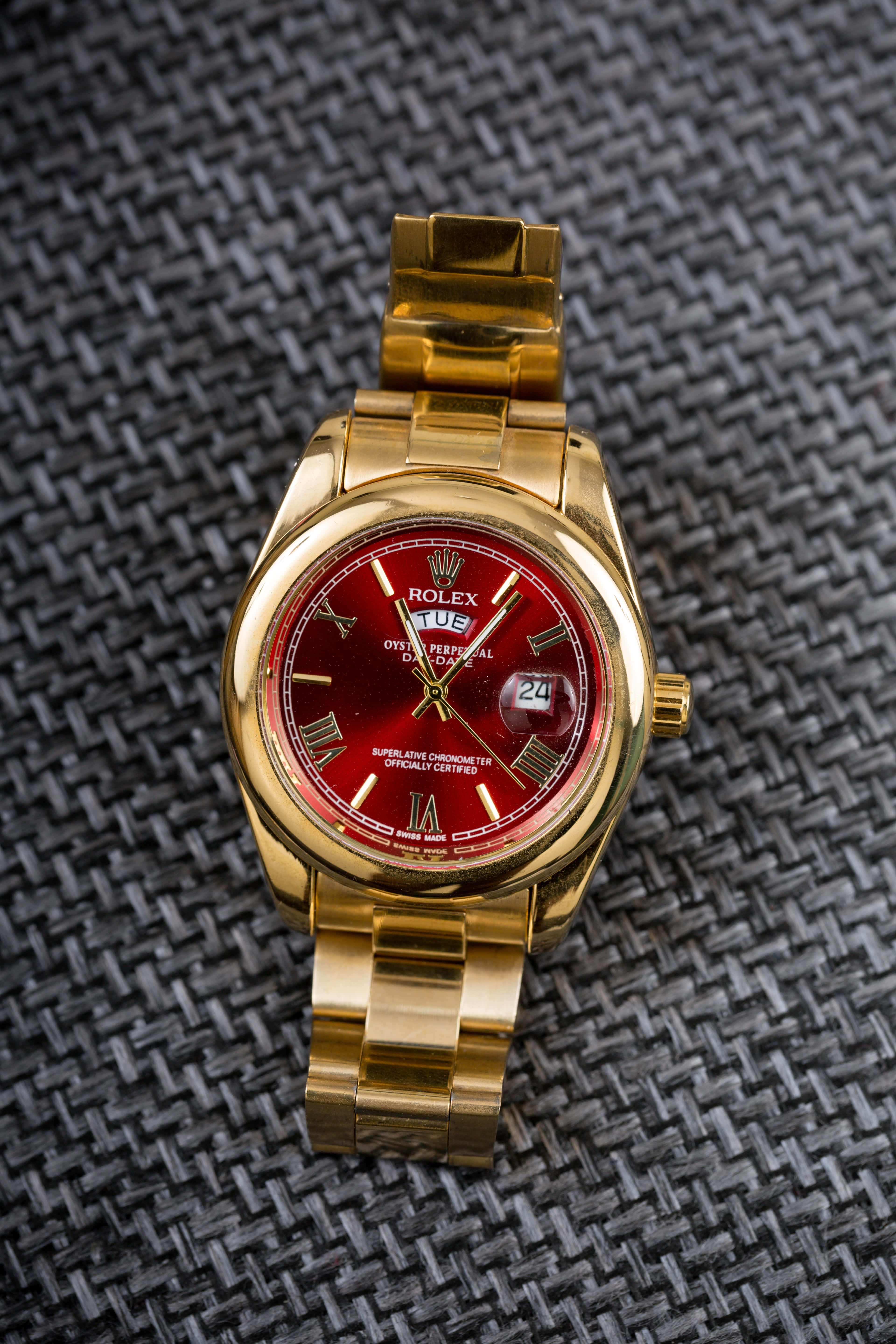 Rolex 桌布 壁紙 — 一隻金色勞力士Day-Date手錶配紅色錶盤，放置在深色編織布料表面上。