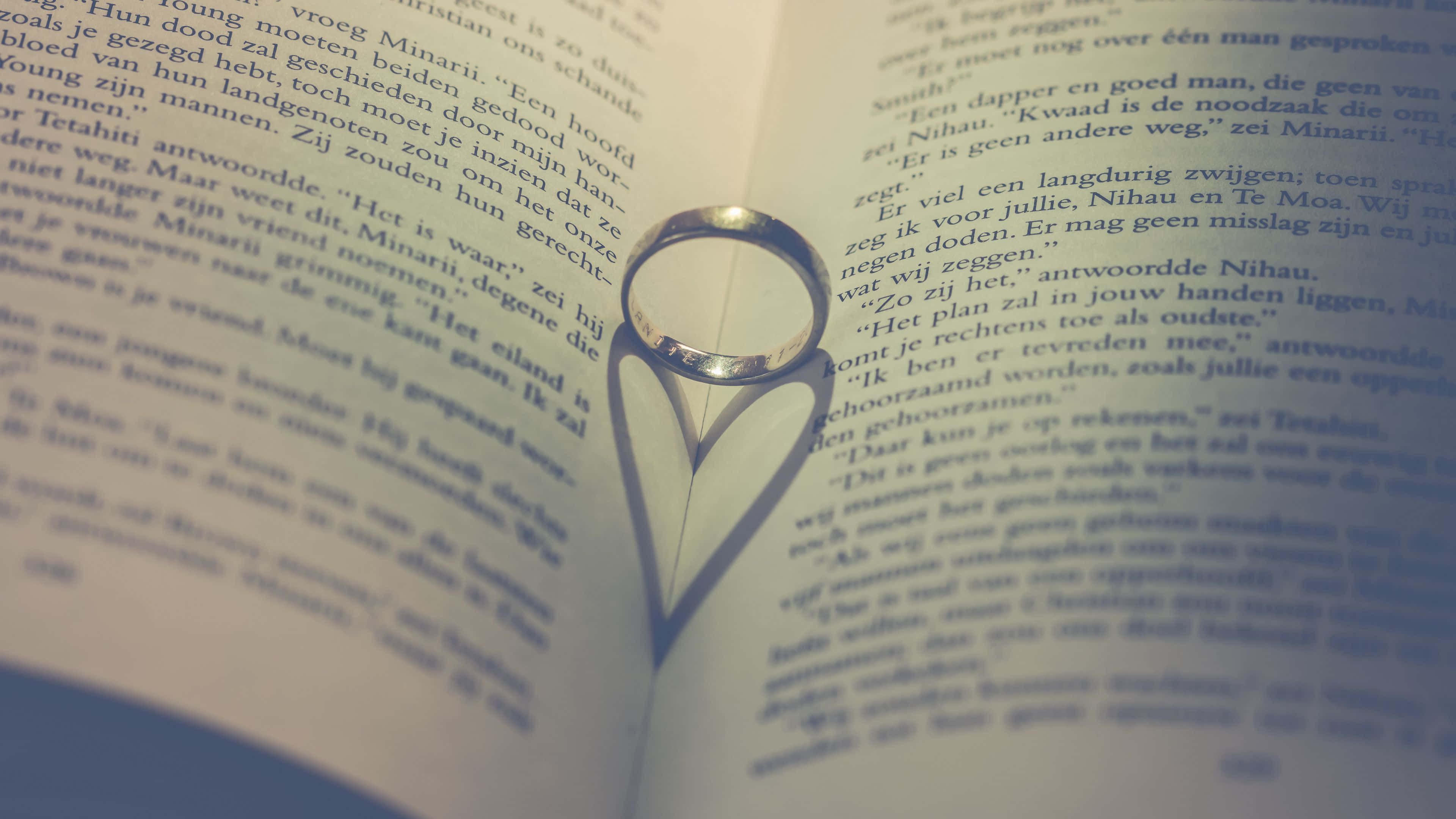 4K Love 桌布 壁紙 — 一隻銀色結婚戒指在柔和燈光下於打開的荷蘭文文字頁面上投射出心形陰影。